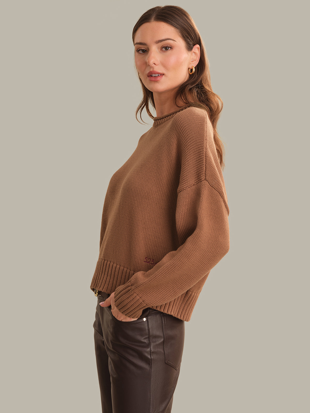 A woman modeling a cropped, long-sleeve ribbed knit sweater in a warm camel brown color, paired with dark brown leather pants, against a solid light gray background.