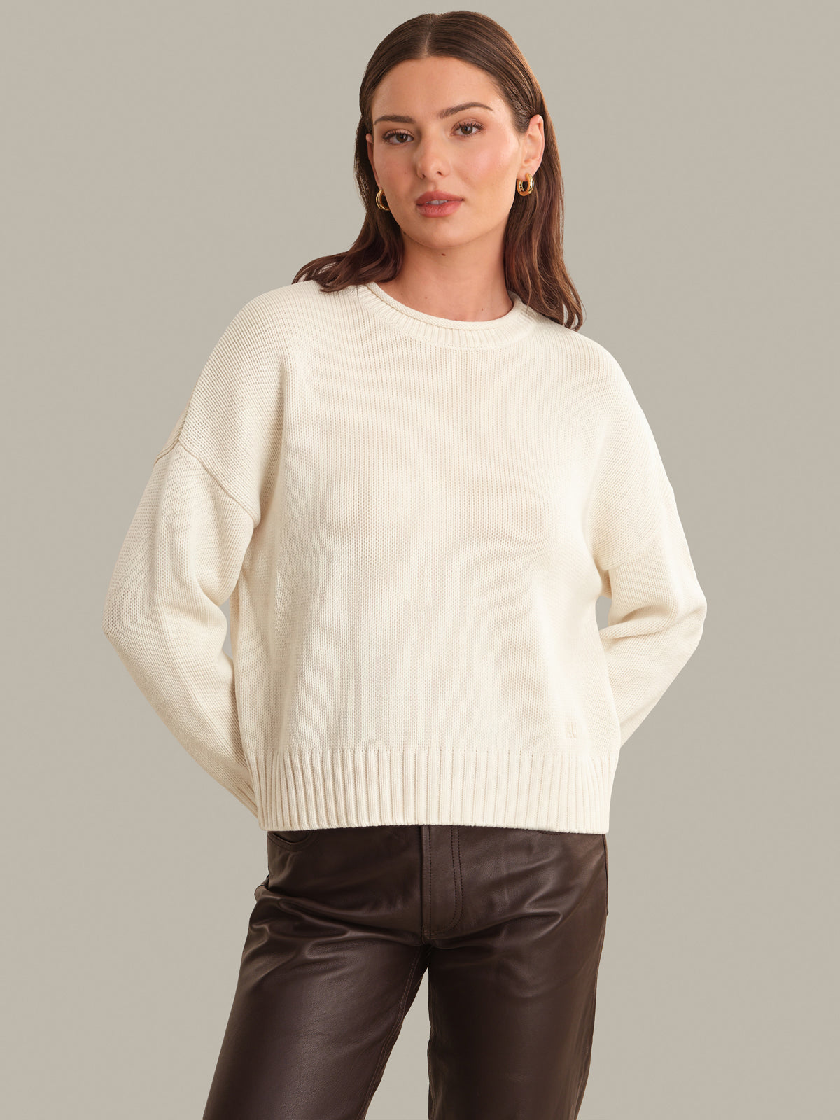 A woman wearing a cream ribbed knit crewneck sweater and dark brown faux-leather pants, standing against a solid light gray studio background.