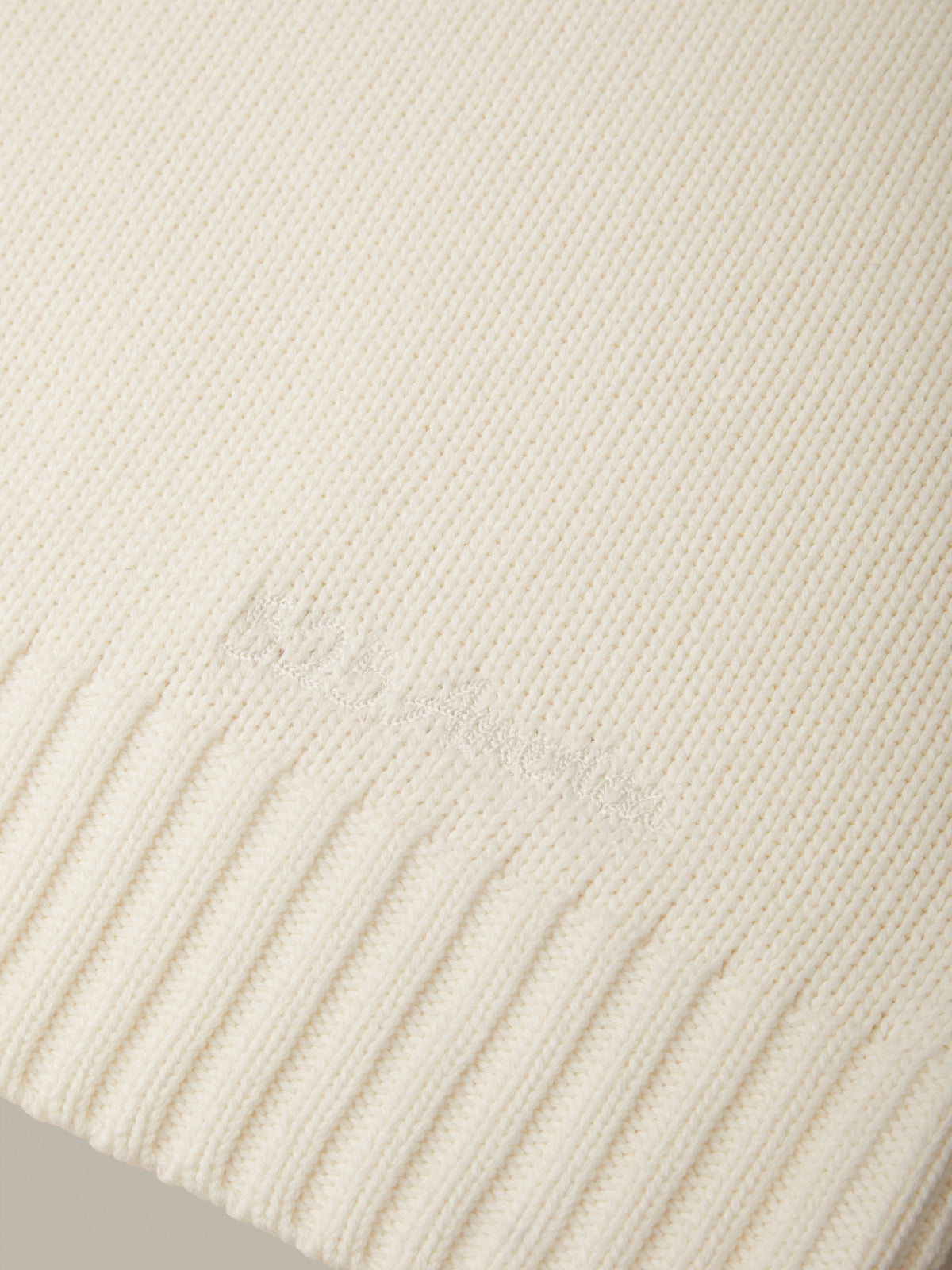 Close-up of a cream-colored ribbed knit fabric with a subtle tonal embroidered logo near the lower left.