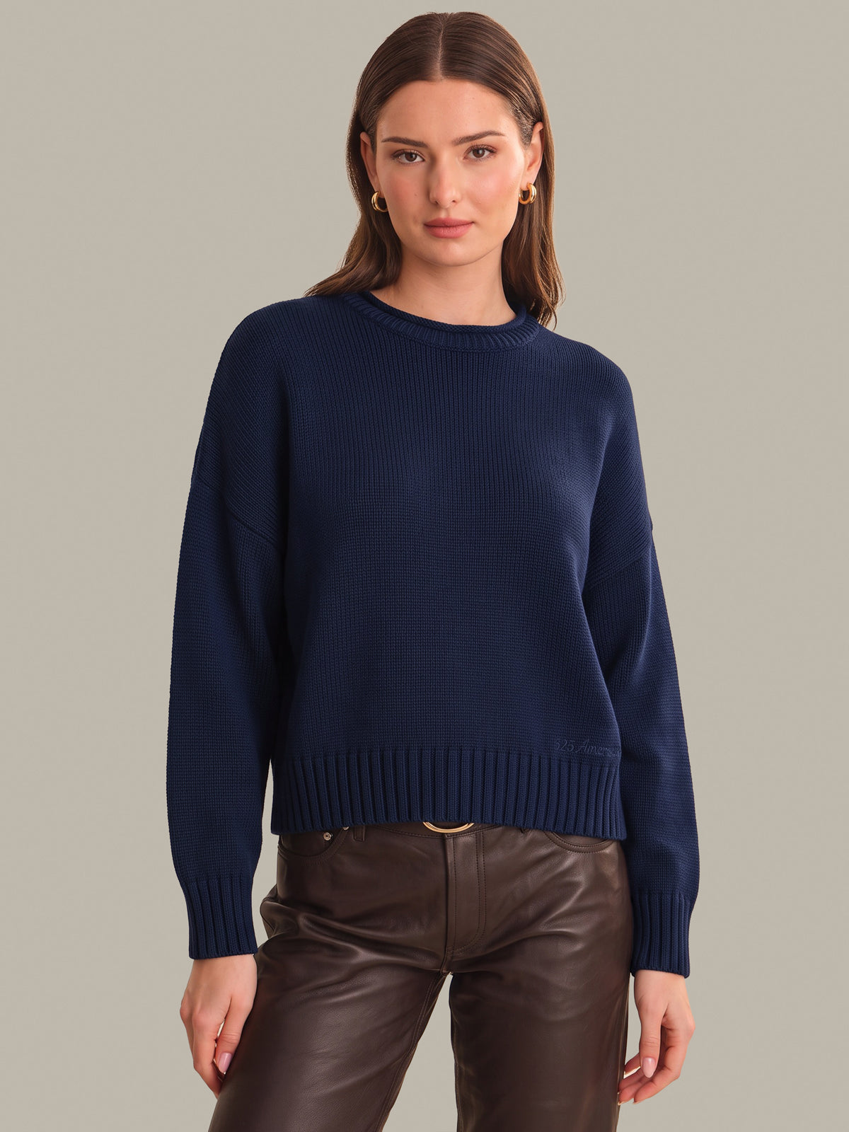 A woman modeling a navy blue ribbed knit sweater with long sleeves and a crew neck, paired with dark brown pants, against a solid light gray background.