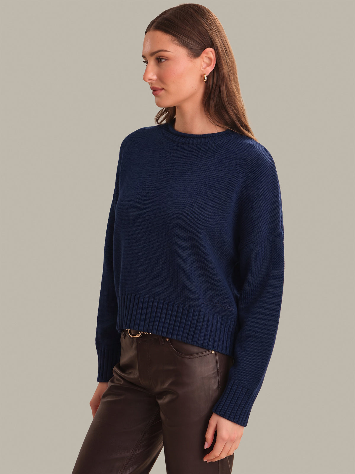 A woman wearing a navy blue knit sweater and dark brown pants, photographed against a plain warm gray studio background.
