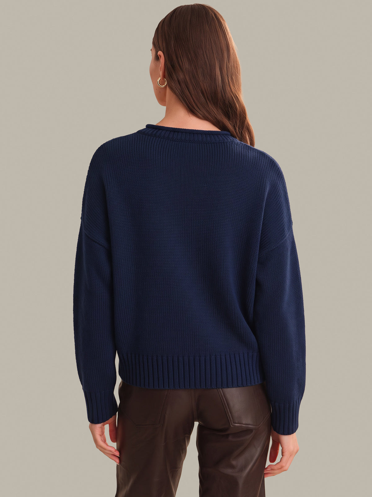 A woman viewed from the back wearing a navy blue ribbed knit sweater and brown pants, standing against a plain light gray studio background.