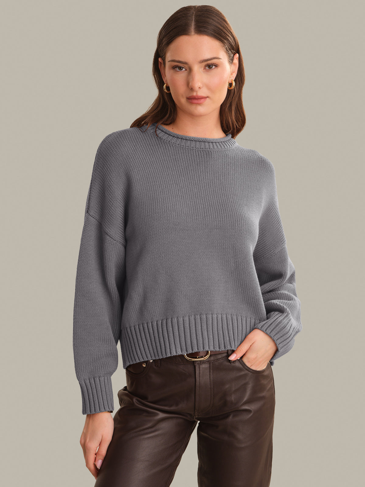 A woman wearing a gray ribbed knit sweater and dark brown pants, standing against a solid light gray background.