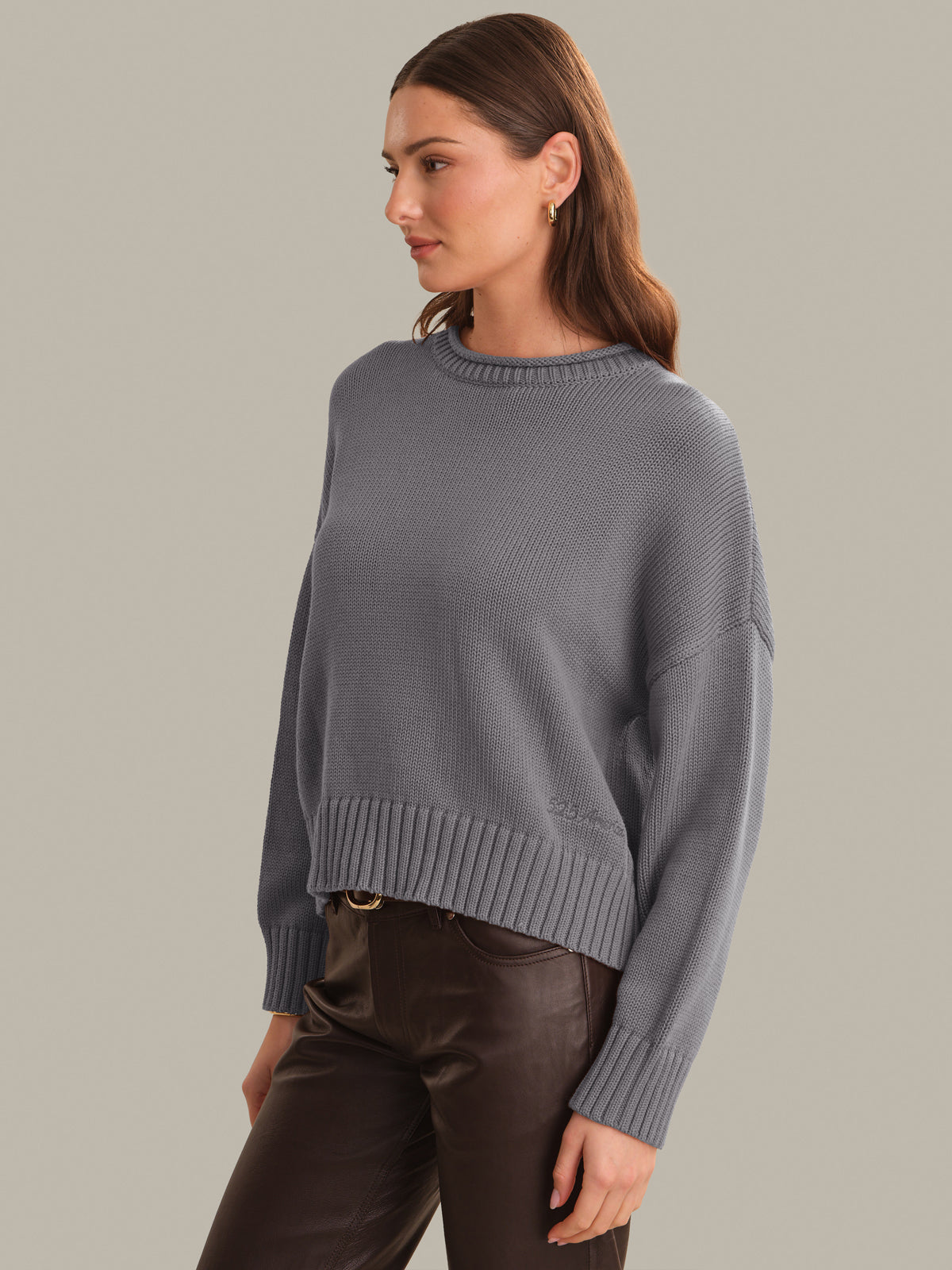 A woman wearing a gray ribbed knit sweater and dark brown pants, shown from the waist up against a solid light gray background.