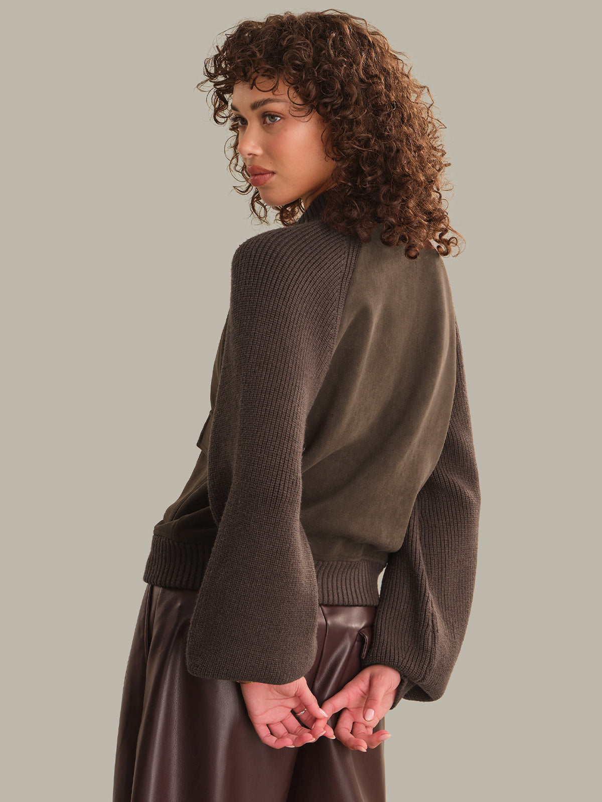 A model with curly brown hair is shown from a three-quarters back view, wearing a dark brown ribbed knit sweater with flared bell sleeves and a dark brown leather skirt, set against a solid light gray background.