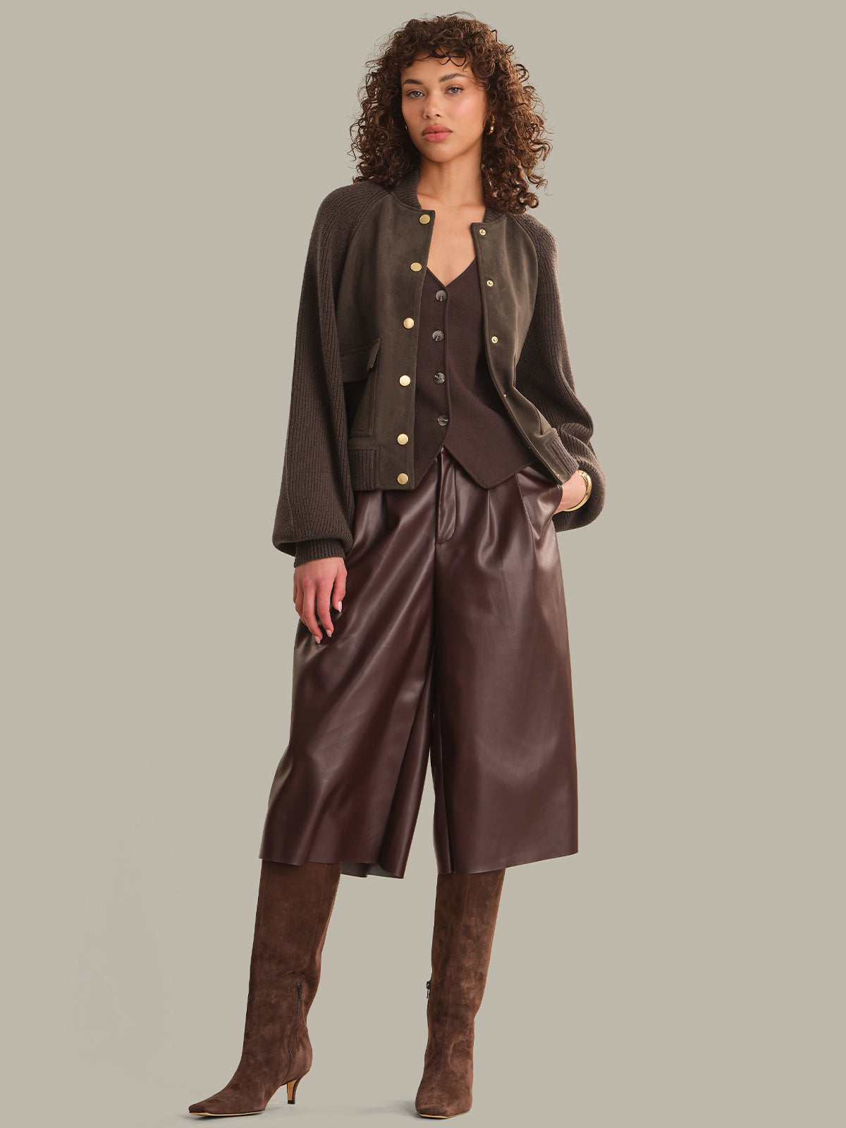 A model wearing a dark brown outfit consisting of a cropped button-front jacket, matching vest, wide-leg faux-leather pants, and tall brown suede boots, set against a solid taupe background.