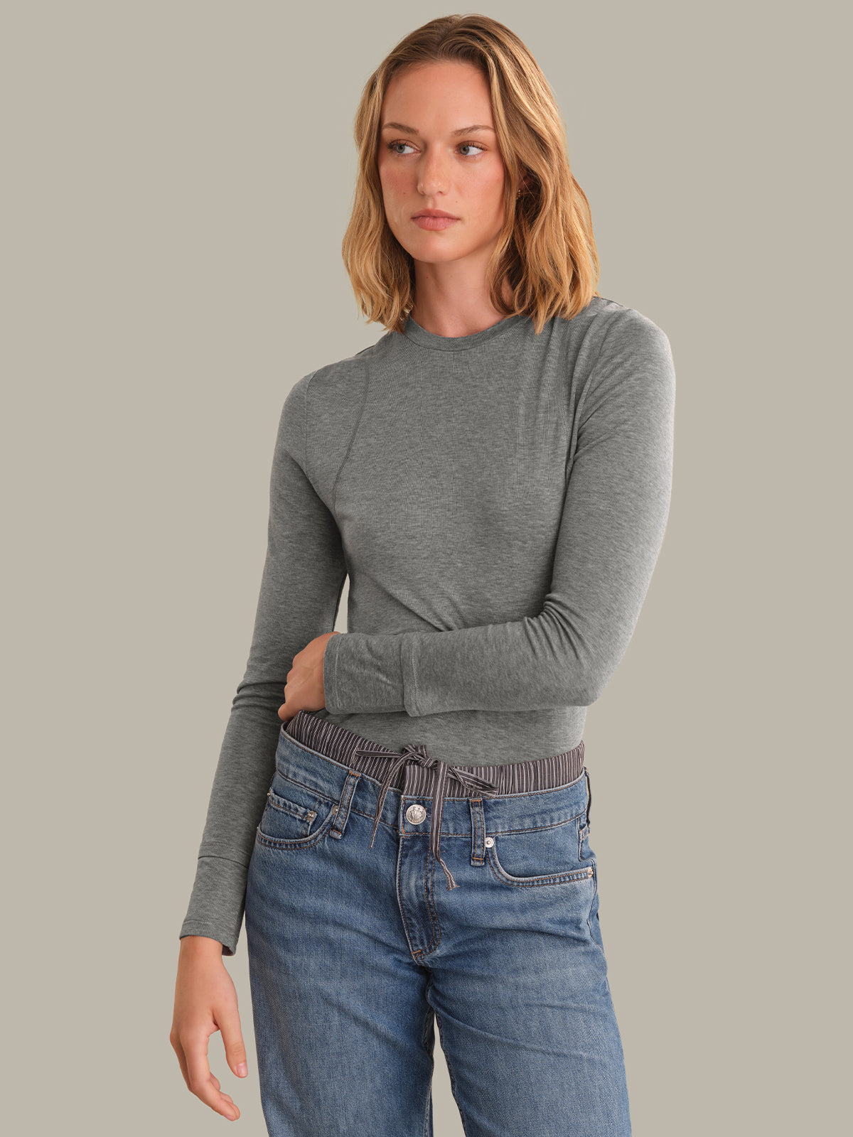 A woman modeling a fitted long-sleeve crewneck top in heather gray, paired with blue jeans, against a solid light gray studio background.