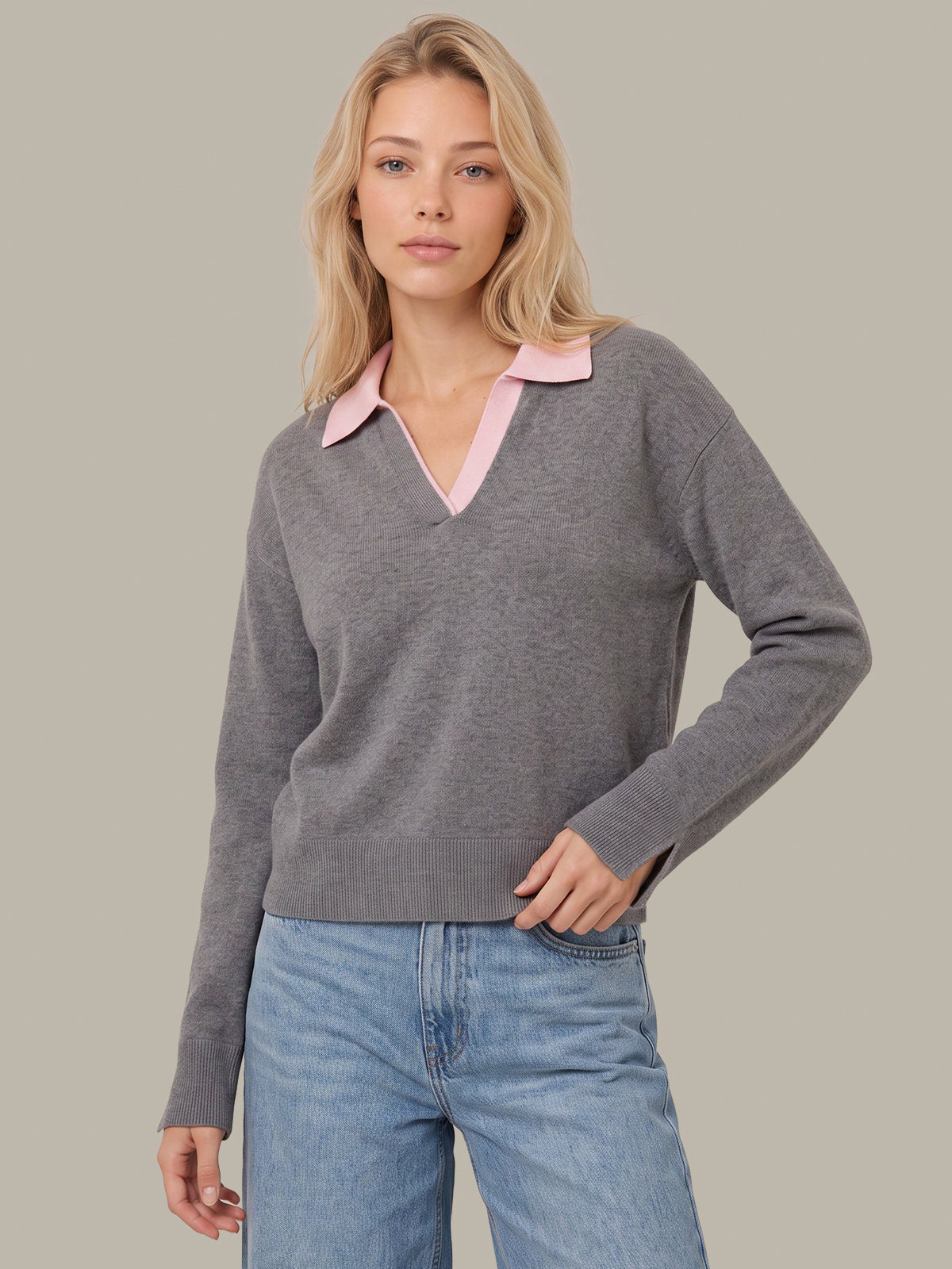A woman modeling a gray long-sleeve knit sweater with a contrasting light pink collar, paired with light blue jeans, against a solid neutral gray background.
