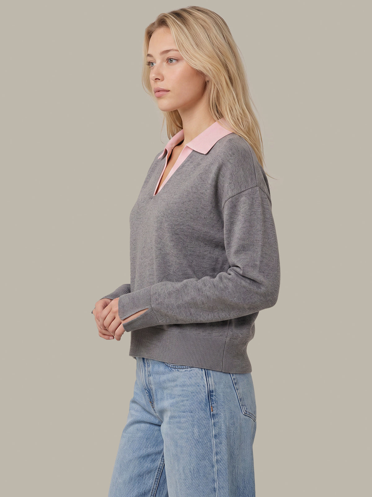 A woman wearing a gray long-sleeve knit sweater with a pink collar, paired with light blue jeans, shown in a side-profile view against a solid beige background.