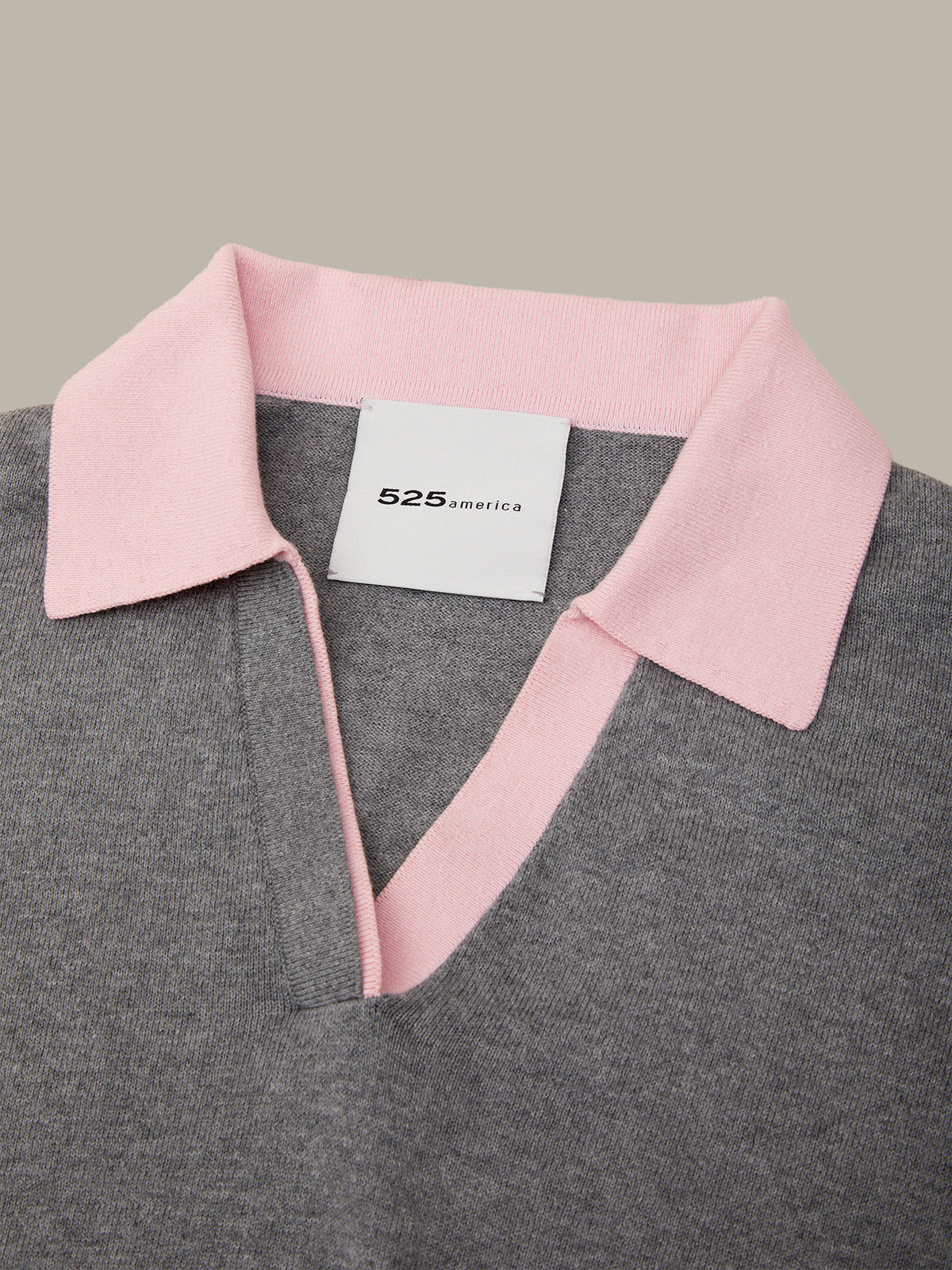 Close-up of a gray knit polo shirt with a light pink collar and a white 525 fashion brand label inside the collar, set against a solid taupe background.