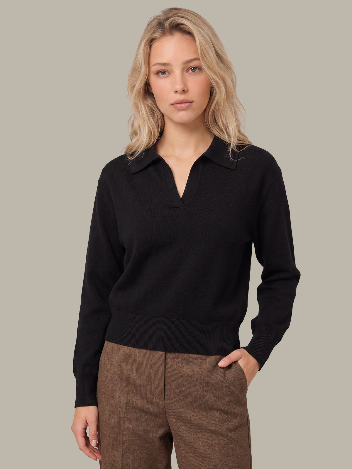 A woman modeling a black long-sleeve knit polo-collar sweater with a split V-neck, paired with brown trousers, against a solid light gray background.