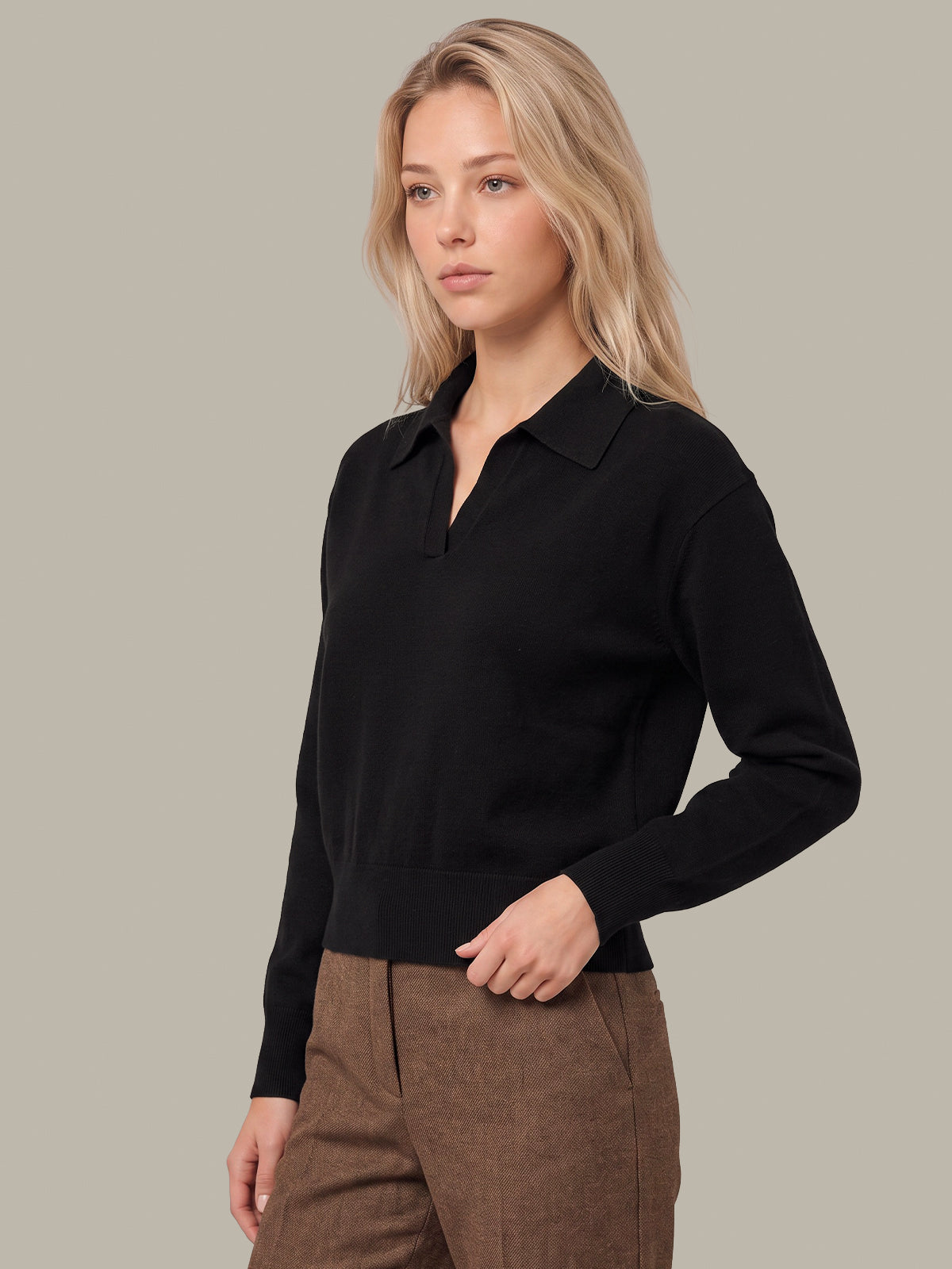 A blonde woman wearing a black long-sleeve polo-knit sweater and brown trousers, standing against a solid light gray studio background.
