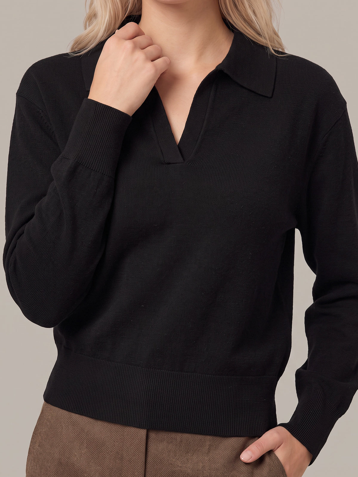 A woman wearing a black long-sleeve knit polo-style sweater with a collar and ribbed hem/cuffs, paired with brown pants, against a plain warm gray studio background.
