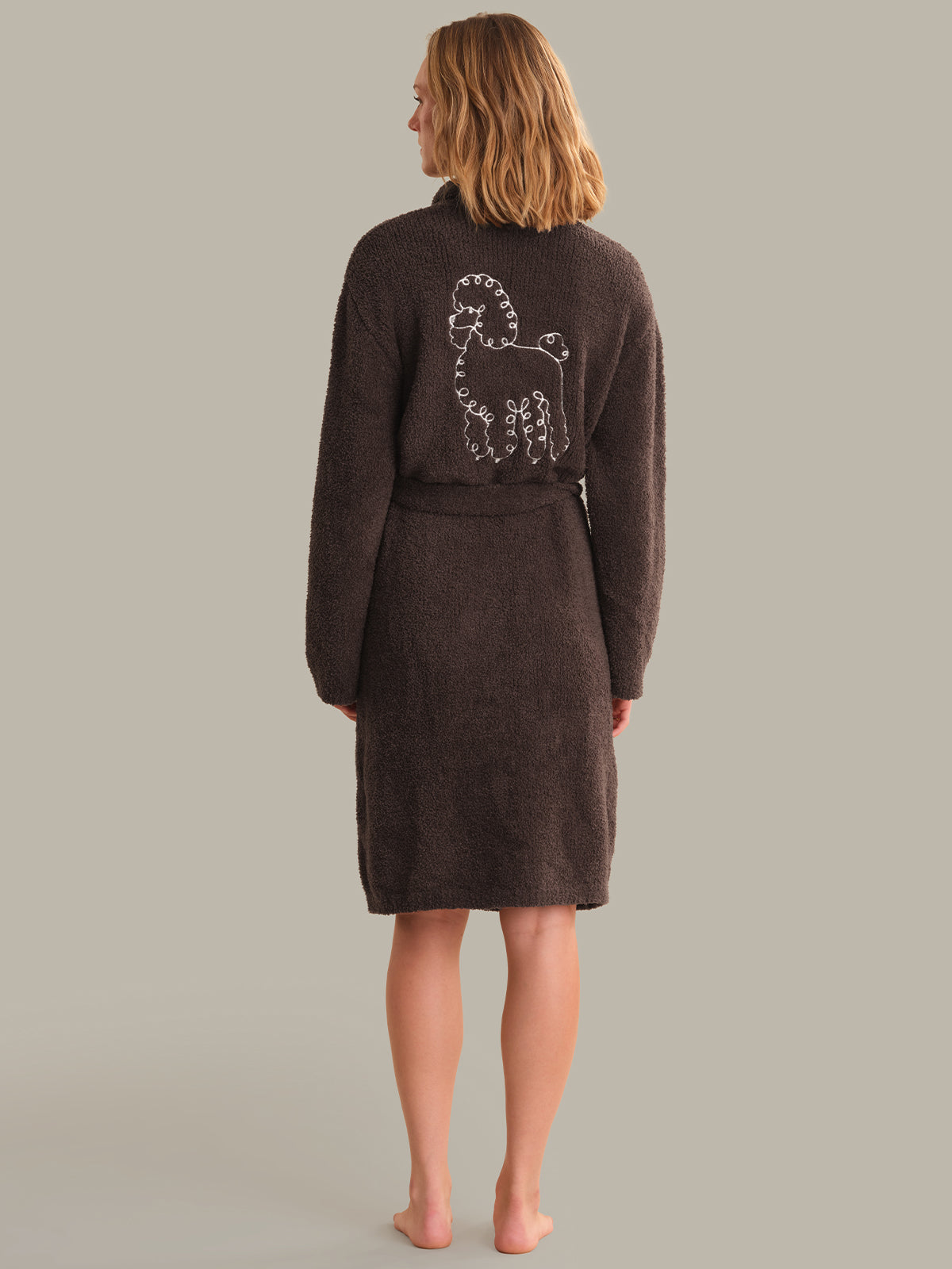A woman viewed from the back wearing a dark brown plush bathrobe with a white poodle outline graphic on the back, standing against a solid light gray background.