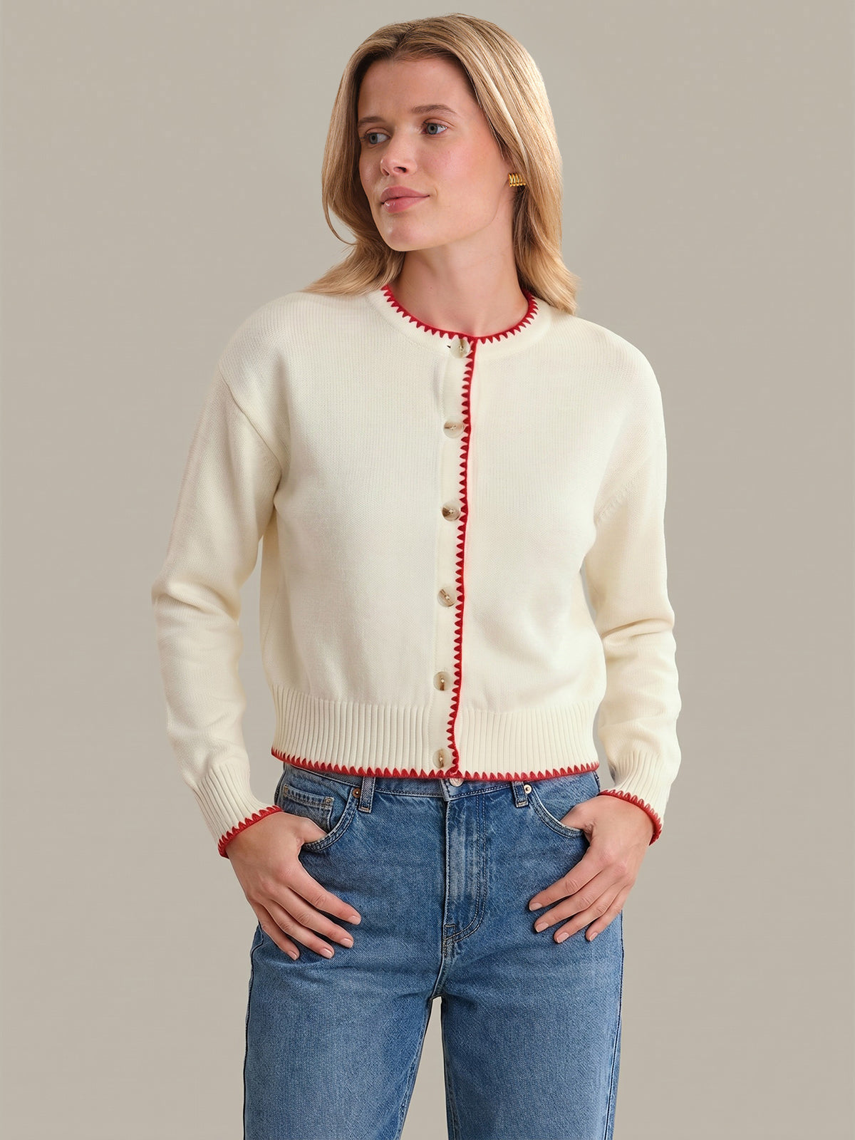Woman wearing a cream cardigan with red trim and blue jeans on a gray background