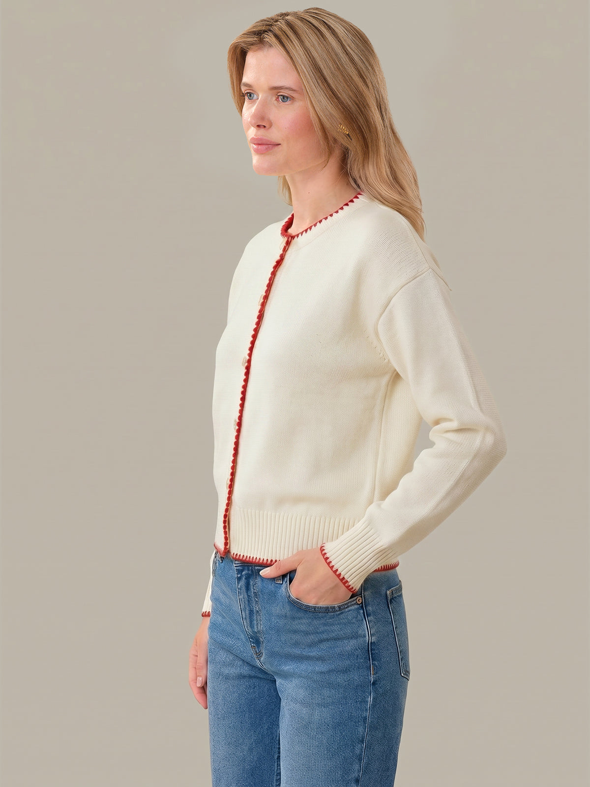 Woman wearing a cream sweater with red trim and blue jeans on a plain background