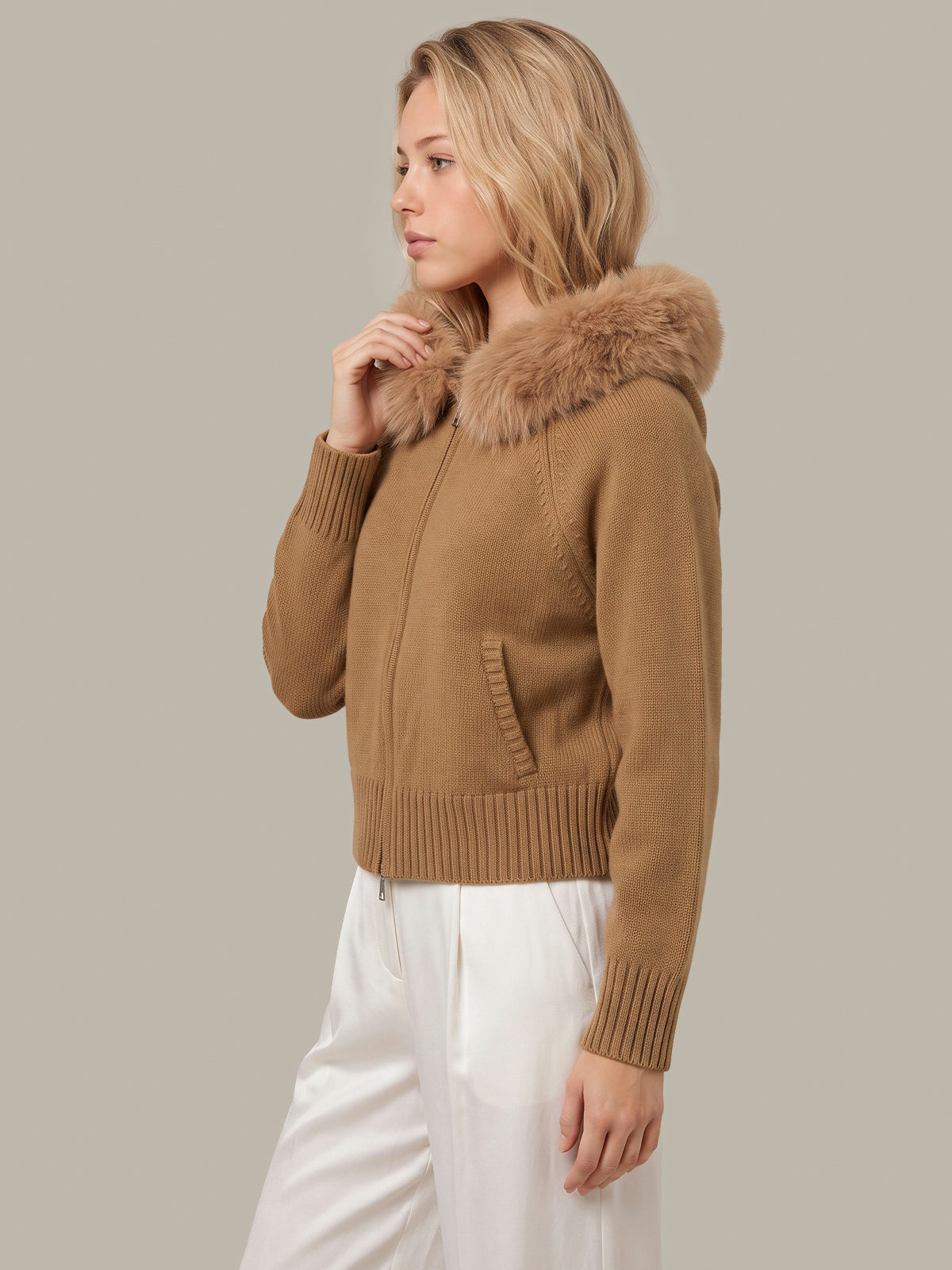A woman wearing a cropped tan knit jacket with a large brown faux-fur collar, paired with white pants, photographed against a solid light gray studio background.