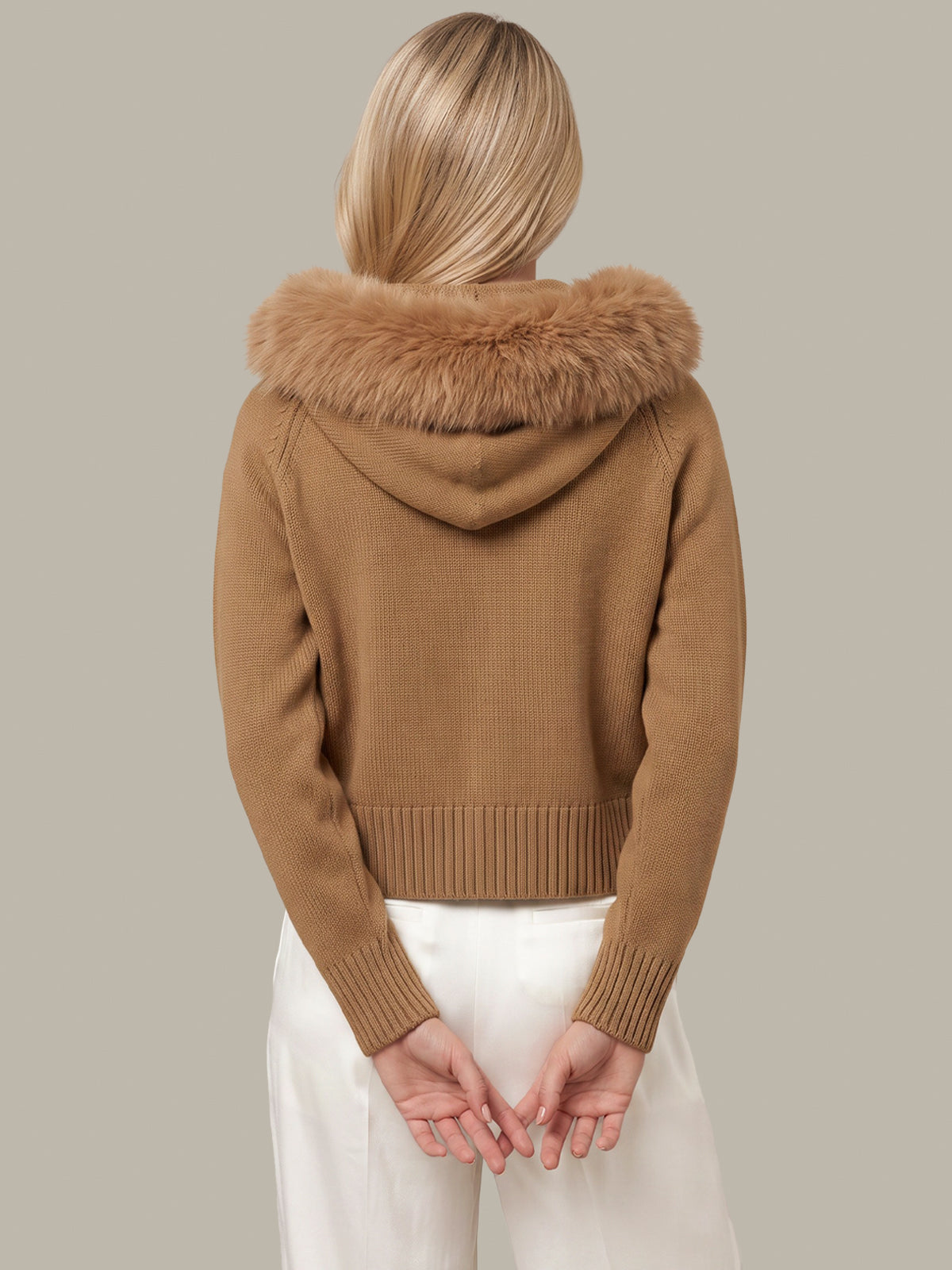 A woman shown from the back wearing a cropped camel brown knit hooded sweater with a fluffy faux-fur trimmed hood, paired with white pants, against a solid light gray background.