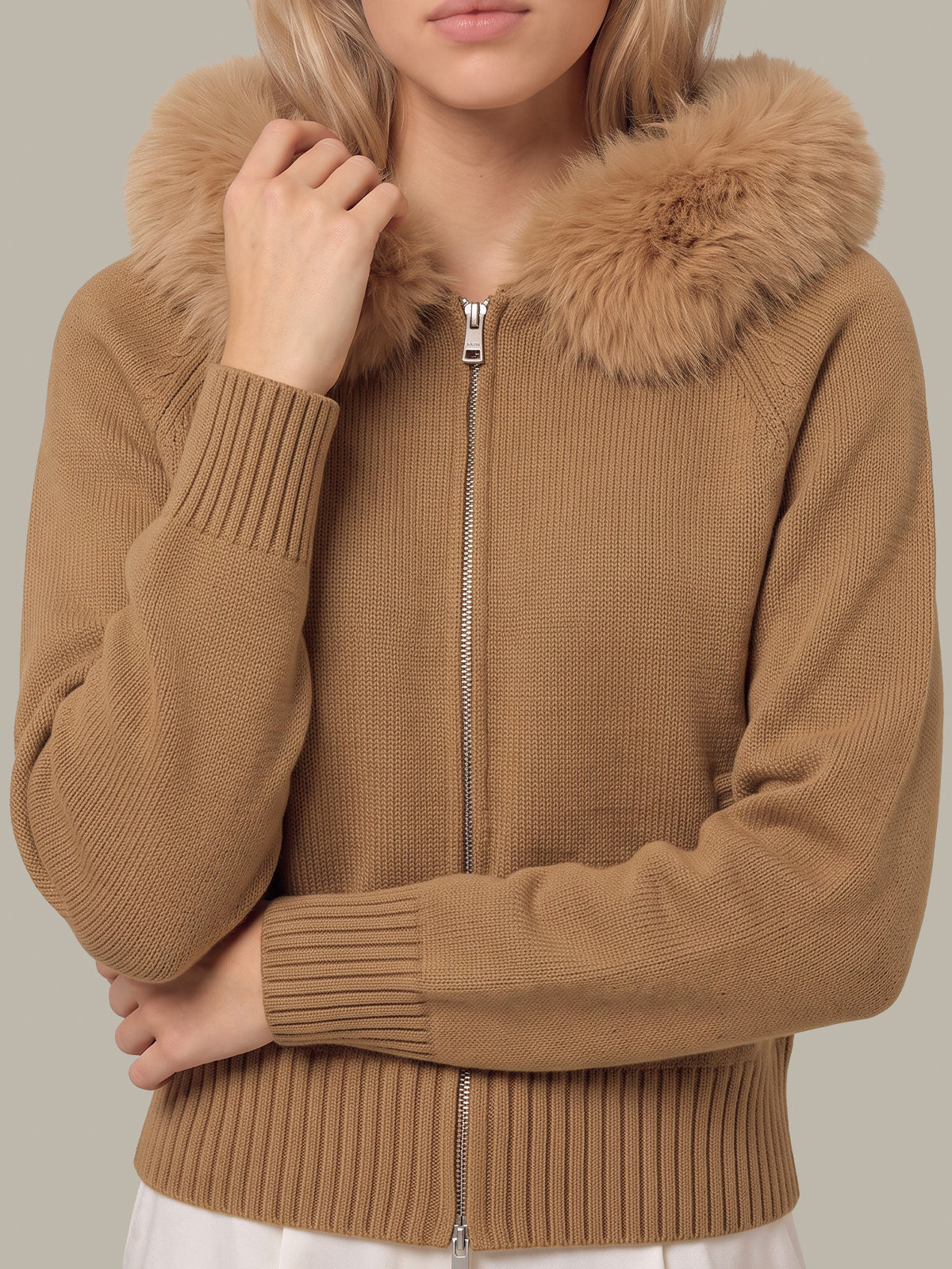 A woman wearing a camel brown ribbed knit zip-up jacket with a large fluffy faux-fur collar, shown from the chin down against a plain light gray studio background.