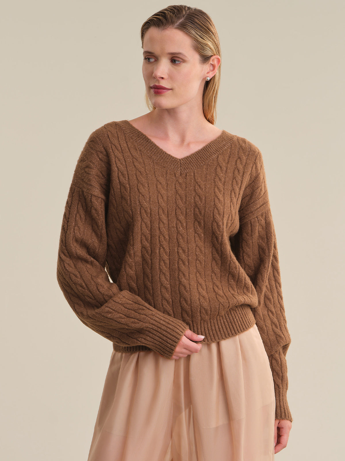 A woman wearing a brown cable-knit V-neck sweater and a light beige skirt, photographed against a plain beige studio background.