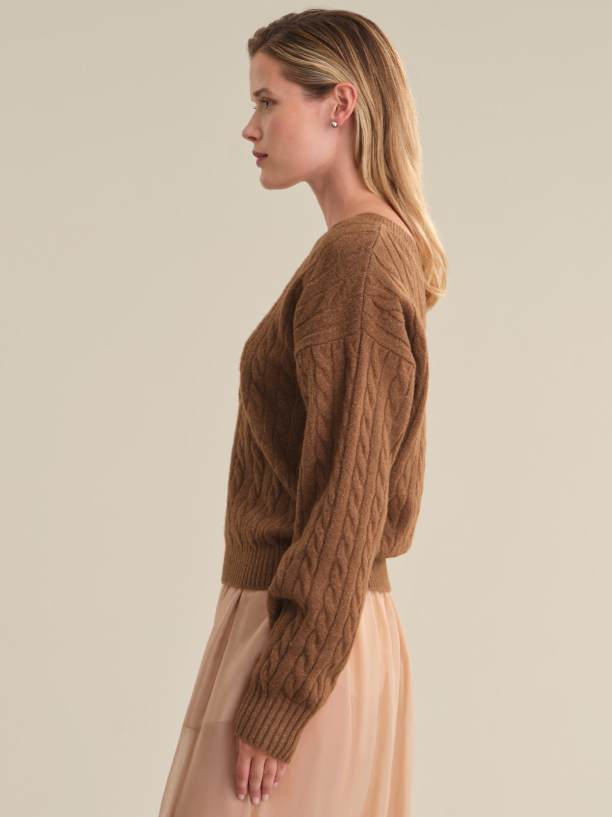 A woman shown in side profile wearing a brown cable-knit sweater and a light peach skirt against a plain beige studio background.