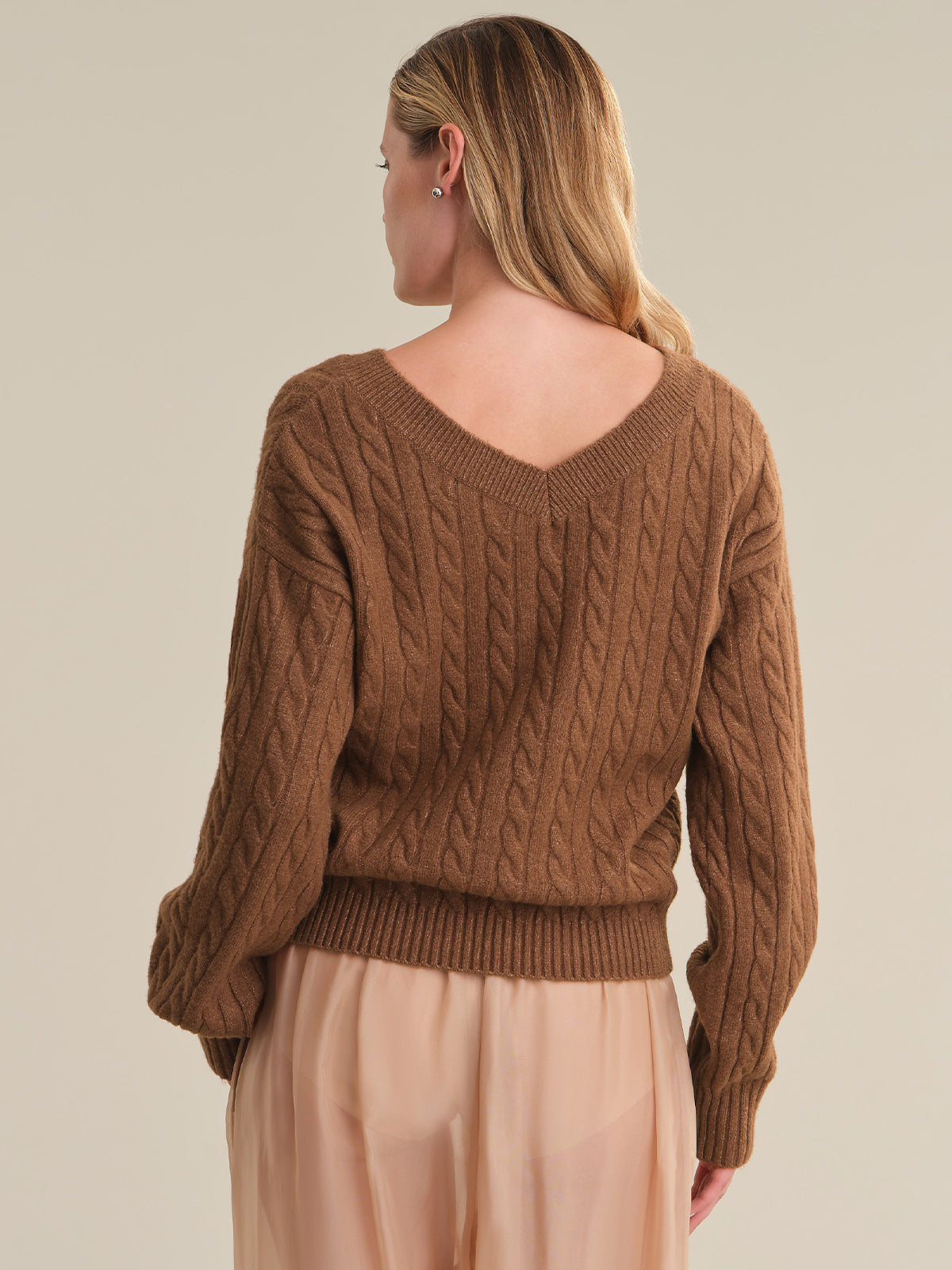 A woman shown from the back wearing a brown cable-knit sweater and a light peach skirt, against a solid beige studio background.
