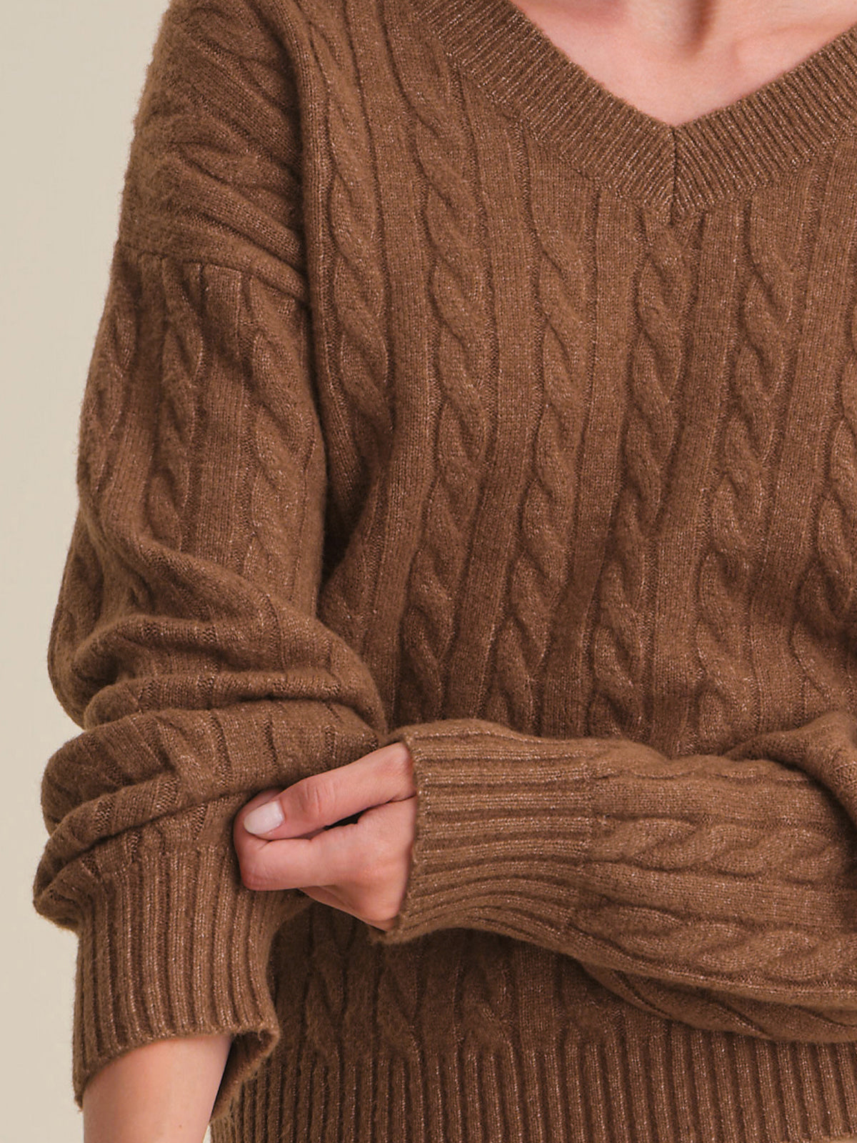 A close-up of a person wearing a brown cable-knit sweater with a V-neck and ribbed cuffs, showing the texture and fit against a plain light beige background.