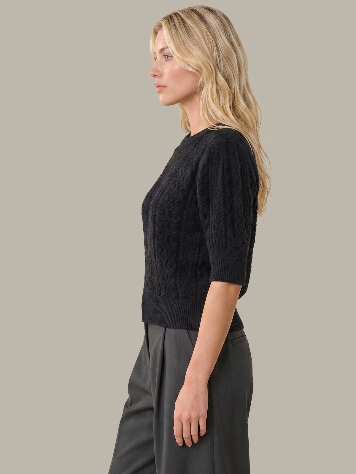 A woman shown in side profile wearing a black textured knit sweater with elbow-length sleeves and dark gray wide-leg trousers, set against a solid warm gray studio background.