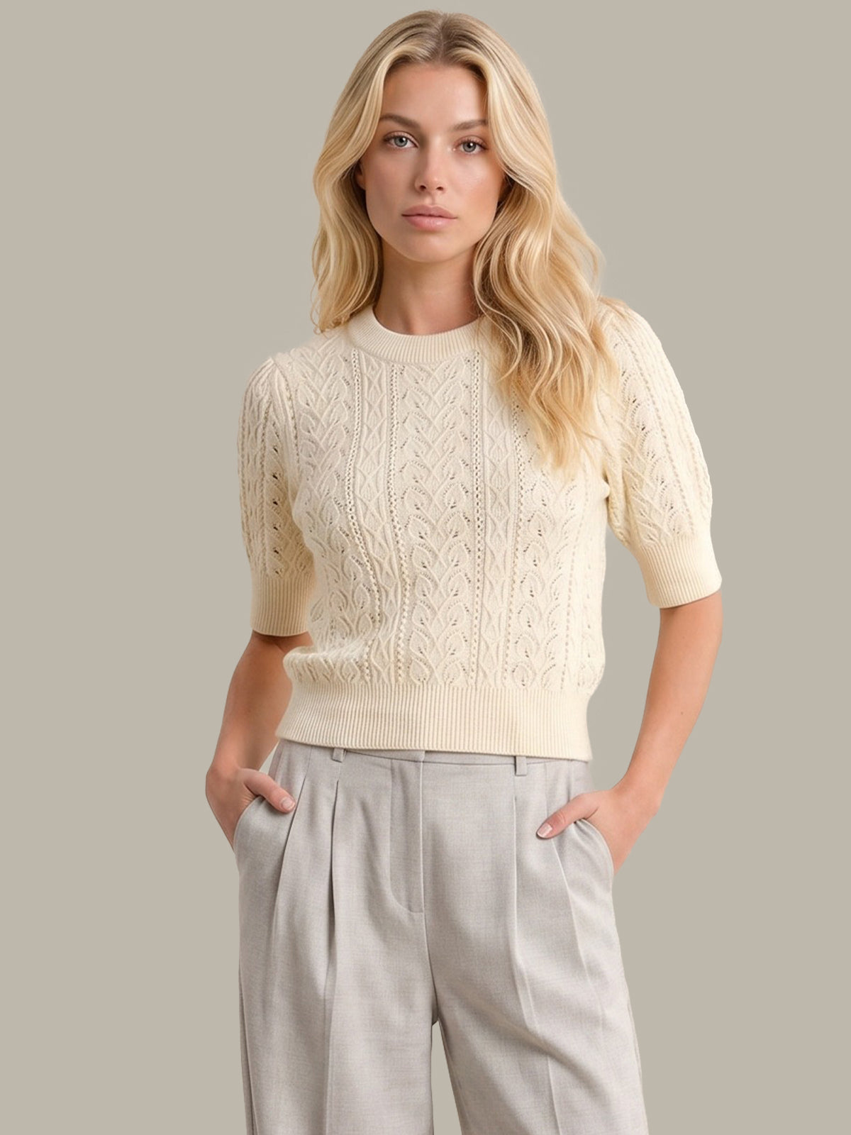 A blonde woman wearing a cream-colored short-sleeve cable-knit sweater and light gray pleated trousers, standing against a solid neutral gray background.