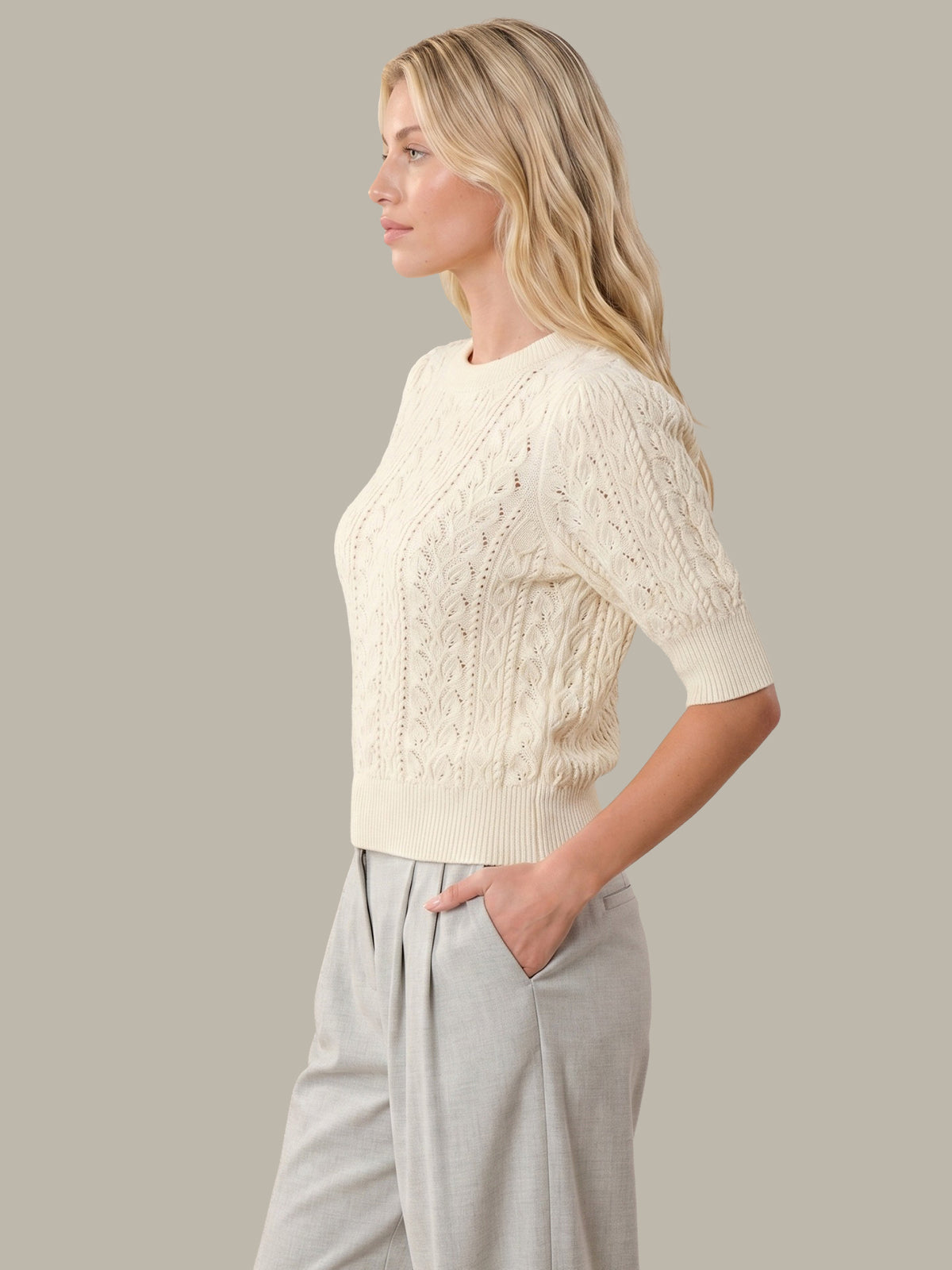 A woman in profile wearing a cream cable-knit short-sleeve sweater and light gray wide-leg pants, photographed against a solid warm gray studio background.