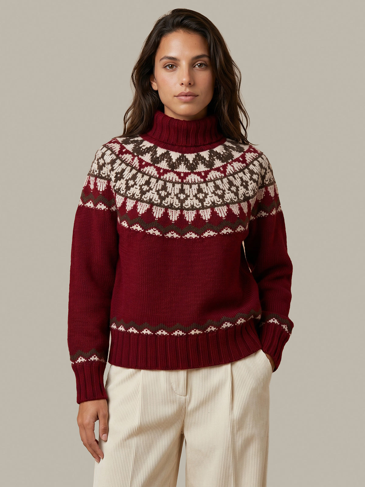A woman wearing a burgundy Fair Isle knit turtleneck sweater with cream and brown patterns, paired with cream trousers, against a solid light gray background.