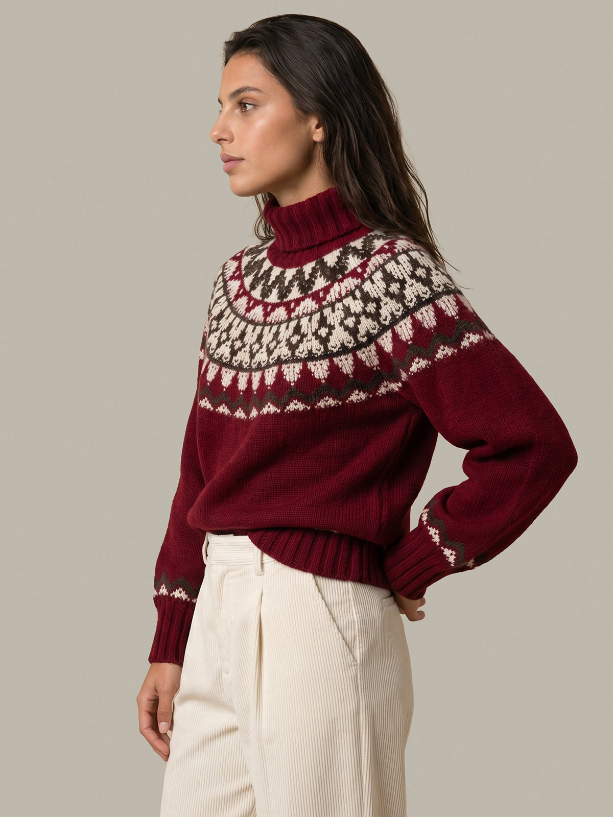 A woman wearing a burgundy turtleneck sweater with a traditional Nordic fair isle pattern in cream and dark brown, paired with cream trousers, set against a solid light gray background.