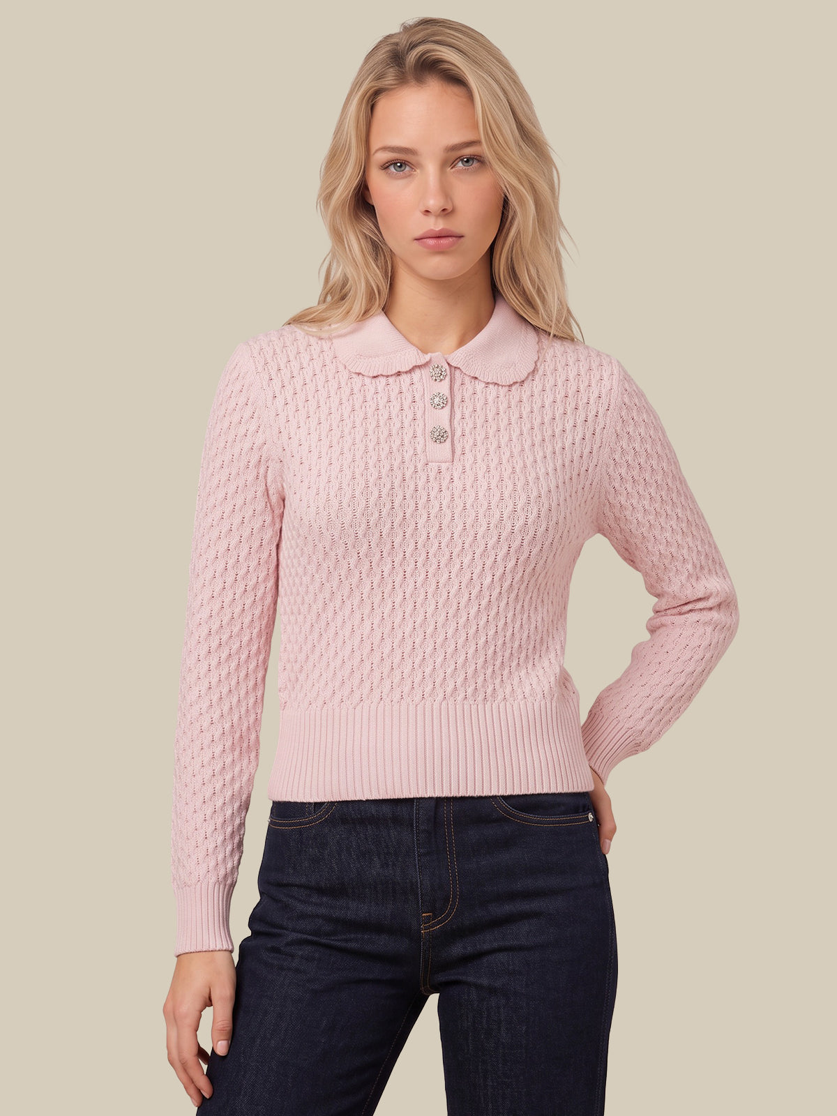 A woman modeling a light pink long-sleeve knit polo sweater with a textured cable pattern and a buttoned collar, paired with dark blue jeans against a solid beige background.