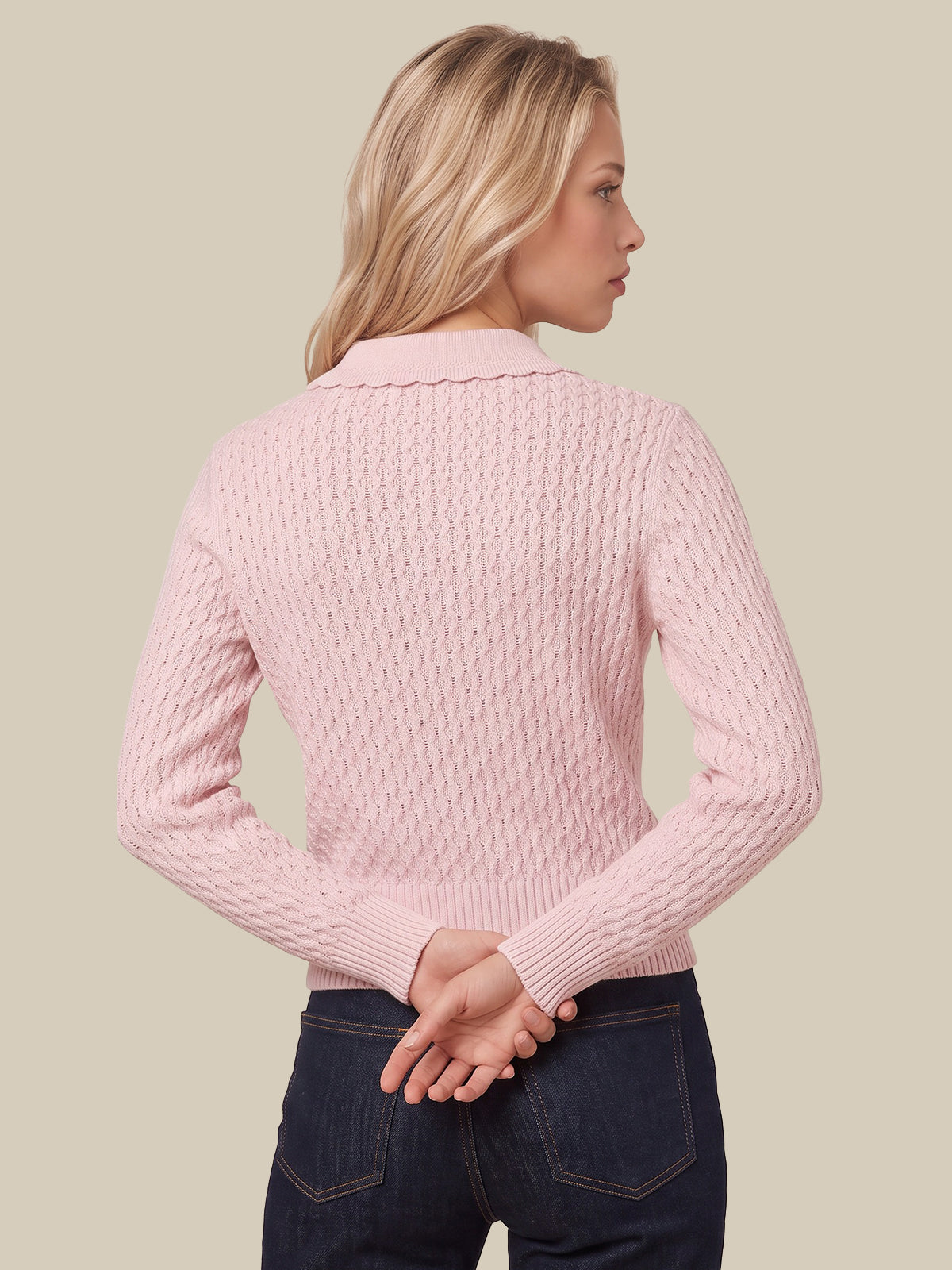 A woman shown from the back wearing a light pink cable-knit sweater with a scalloped collar and dark blue jeans, set against a solid beige background.