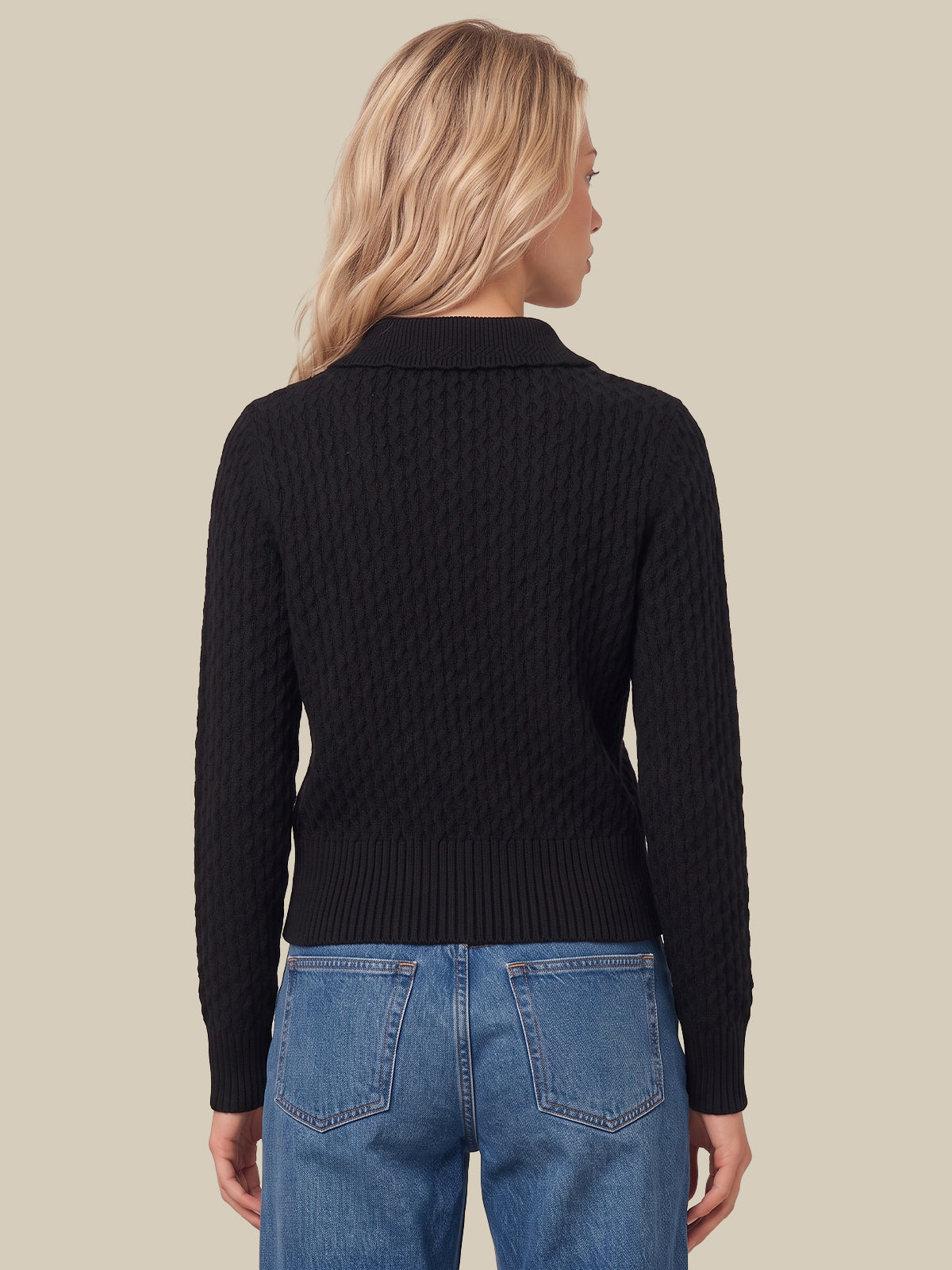 A woman viewed from the back wearing a black cable-knit mock-neck sweater and blue jeans, set against a solid beige background.