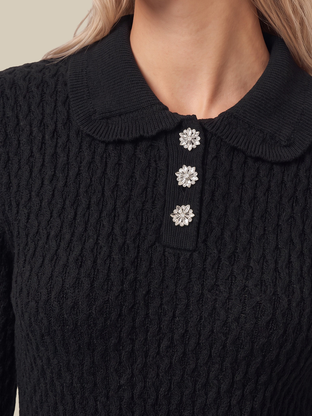 Close-up of a black cable-knit sweater with a Peter Pan collar and three decorative silver flower-shaped buttons on the placket, worn by a model.