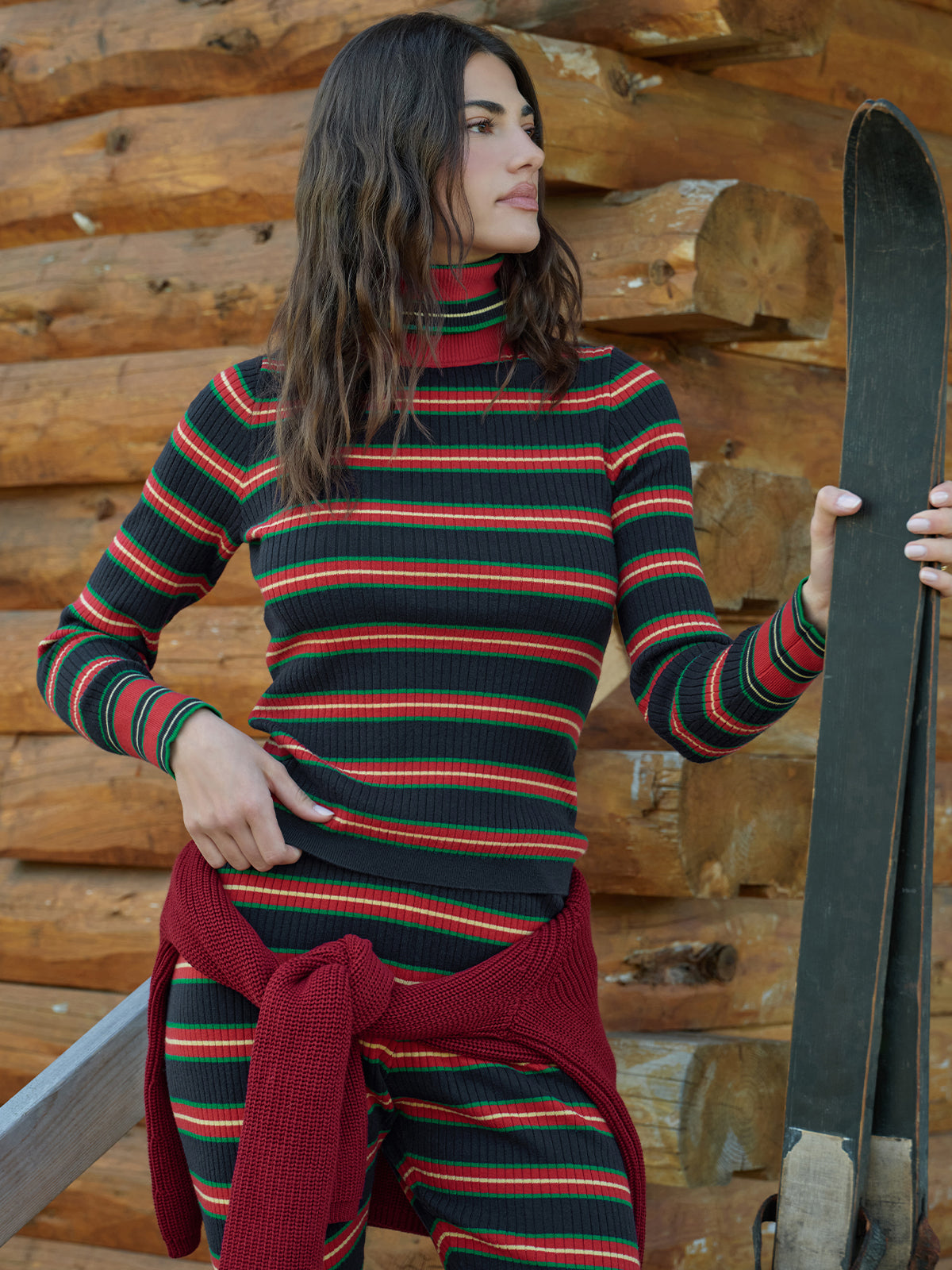 A woman wearing a ribbed turtleneck sweater and matching striped pants, posed in front of a rustic log cabin wall while holding a pair of skis.
