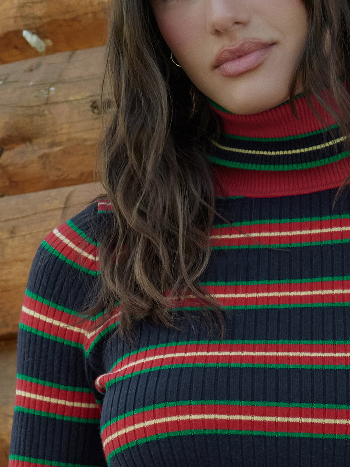 A close-up of a woman wearing a ribbed turtleneck sweater with horizontal stripes in red, green, cream, and navy blue, posed against a rustic wooden background.