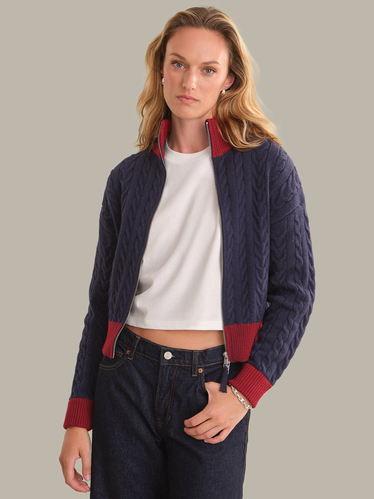 A woman modeling a navy blue cable-knit cropped jacket with contrasting red ribbed trim on the collar, cuffs, and hem, worn over a white crop top and dark jeans against a solid gray background.