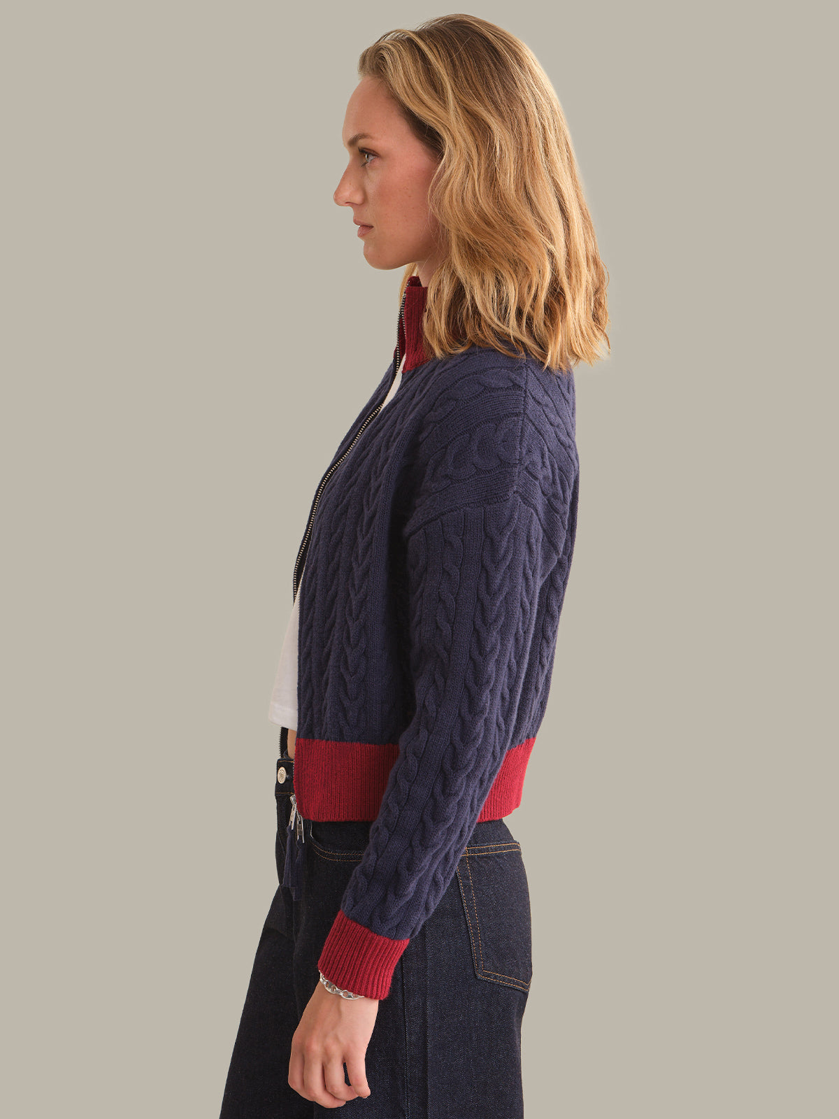 A woman shown in side profile wearing a navy blue cable-knit cardigan with red ribbed trim at the collar, cuffs, and hem, paired with dark jeans and a white top, against a solid light gray background.
