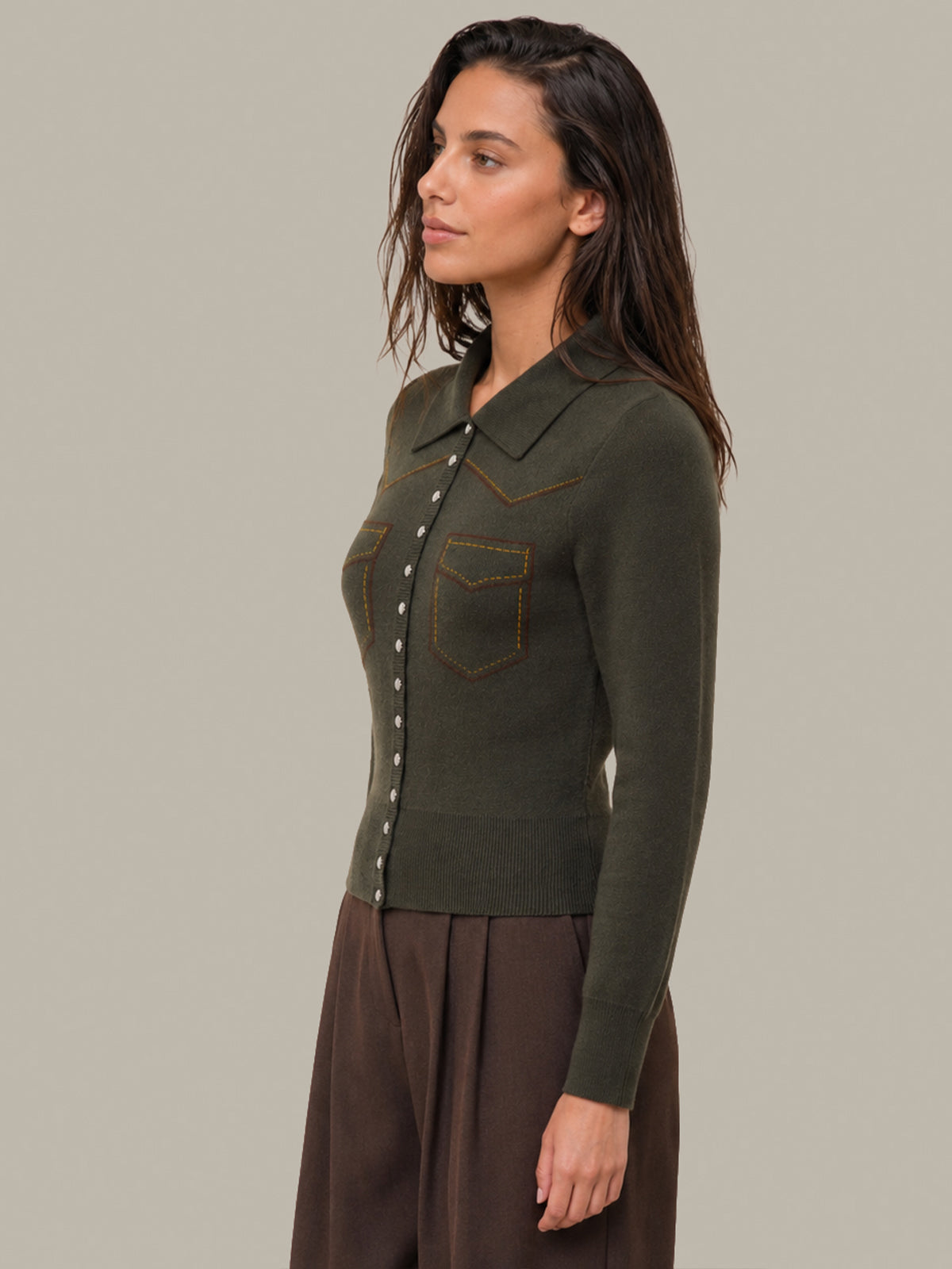 A woman wearing an olive green long-sleeve knit cardigan with western-style chest pockets and brown pants, photographed against a solid light gray studio background.