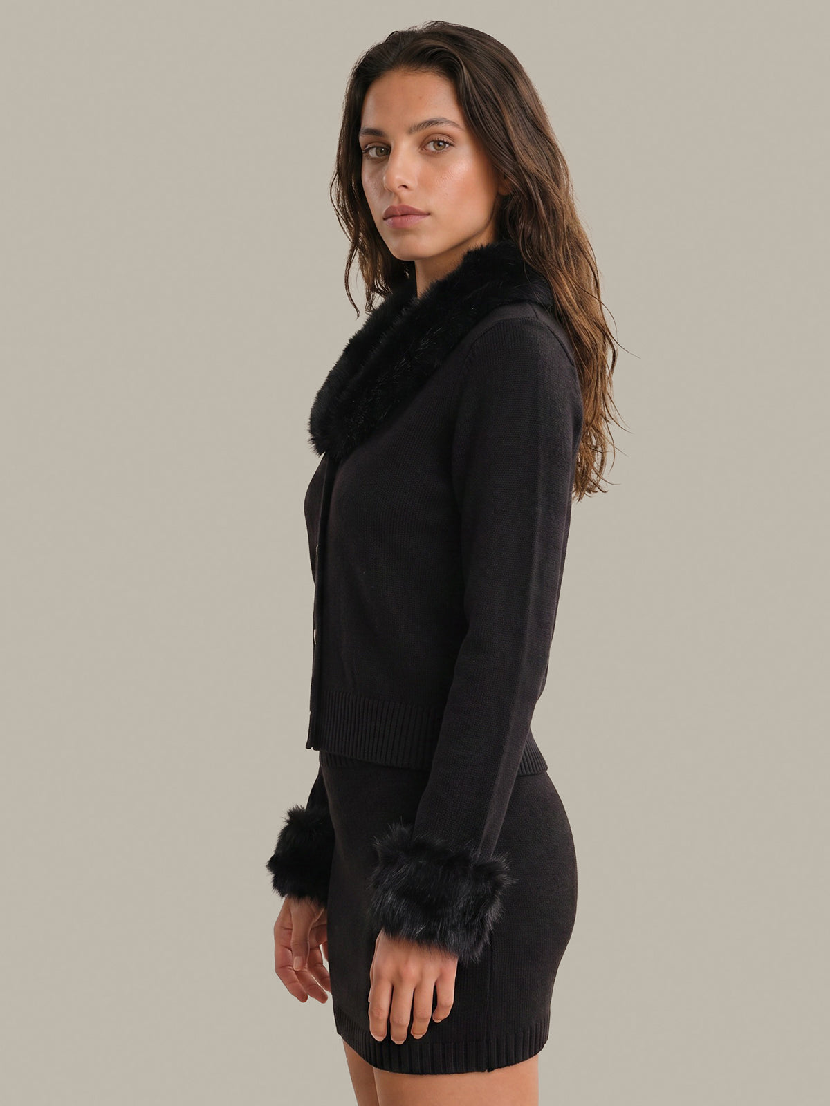 A woman modeling a black knit cardigan with a black faux-fur collar and matching faux-fur trimmed cuffs, paired with a black ribbed mini skirt, against a solid light gray studio background.