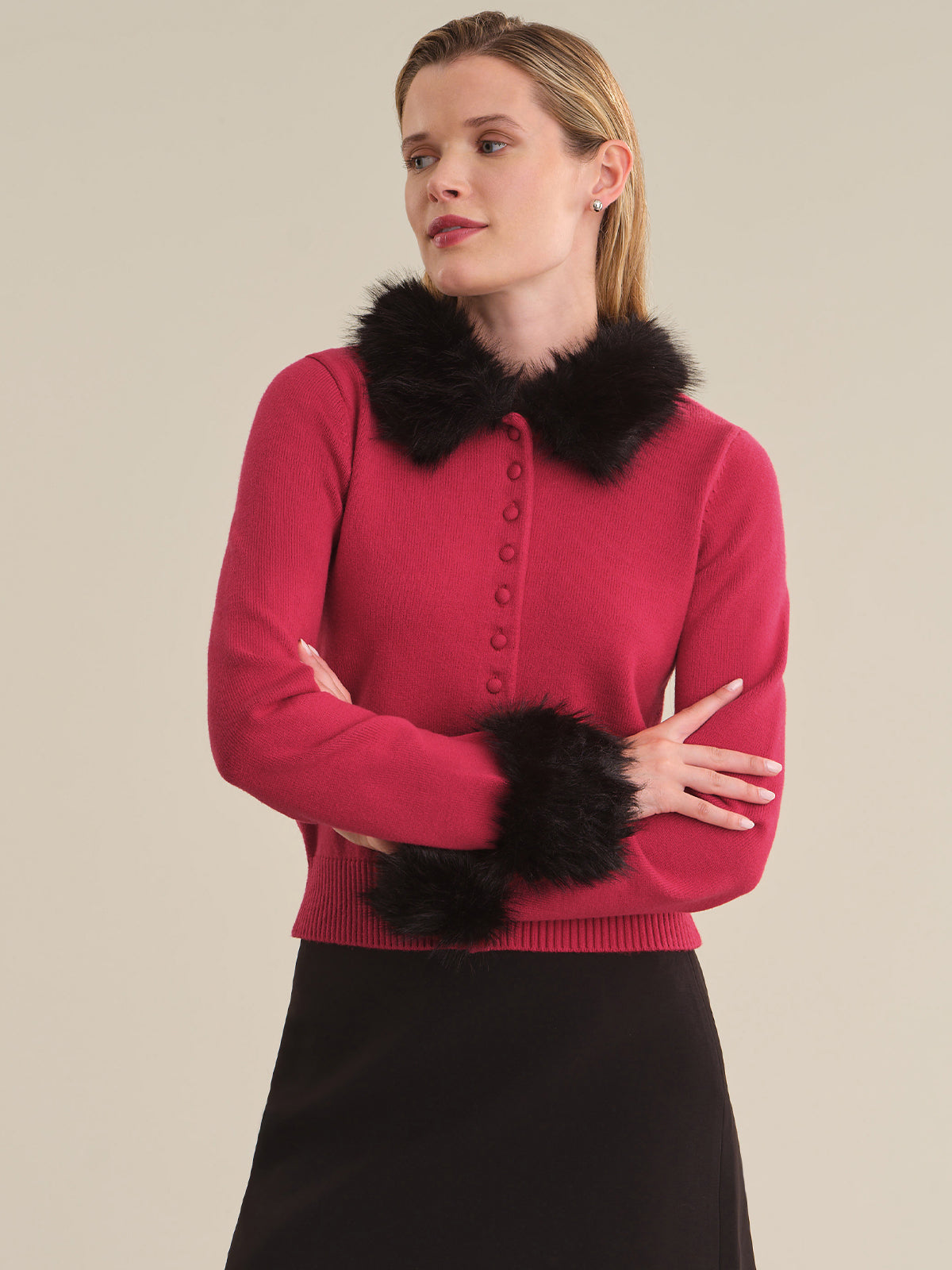 A woman wearing a bright pink knit cardigan with black faux-fur trim at the collar and cuffs, paired with a black skirt, against a solid beige studio background.