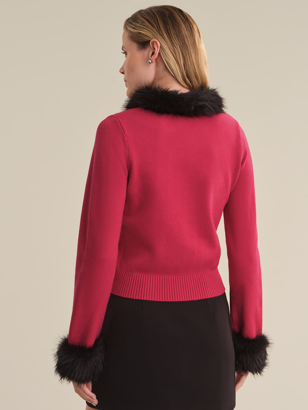 A woman shown from the back wearing a bright pink knit sweater with black faux-fur trim at the collar and cuffs, paired with a black skirt, against a solid beige background.