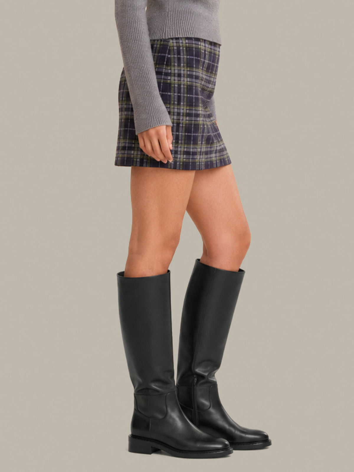 A model wearing a gray ribbed sweater, a dark blue and gray plaid pleated mini skirt, and black leather knee-high boots, shown from the waist down against a solid beige background.