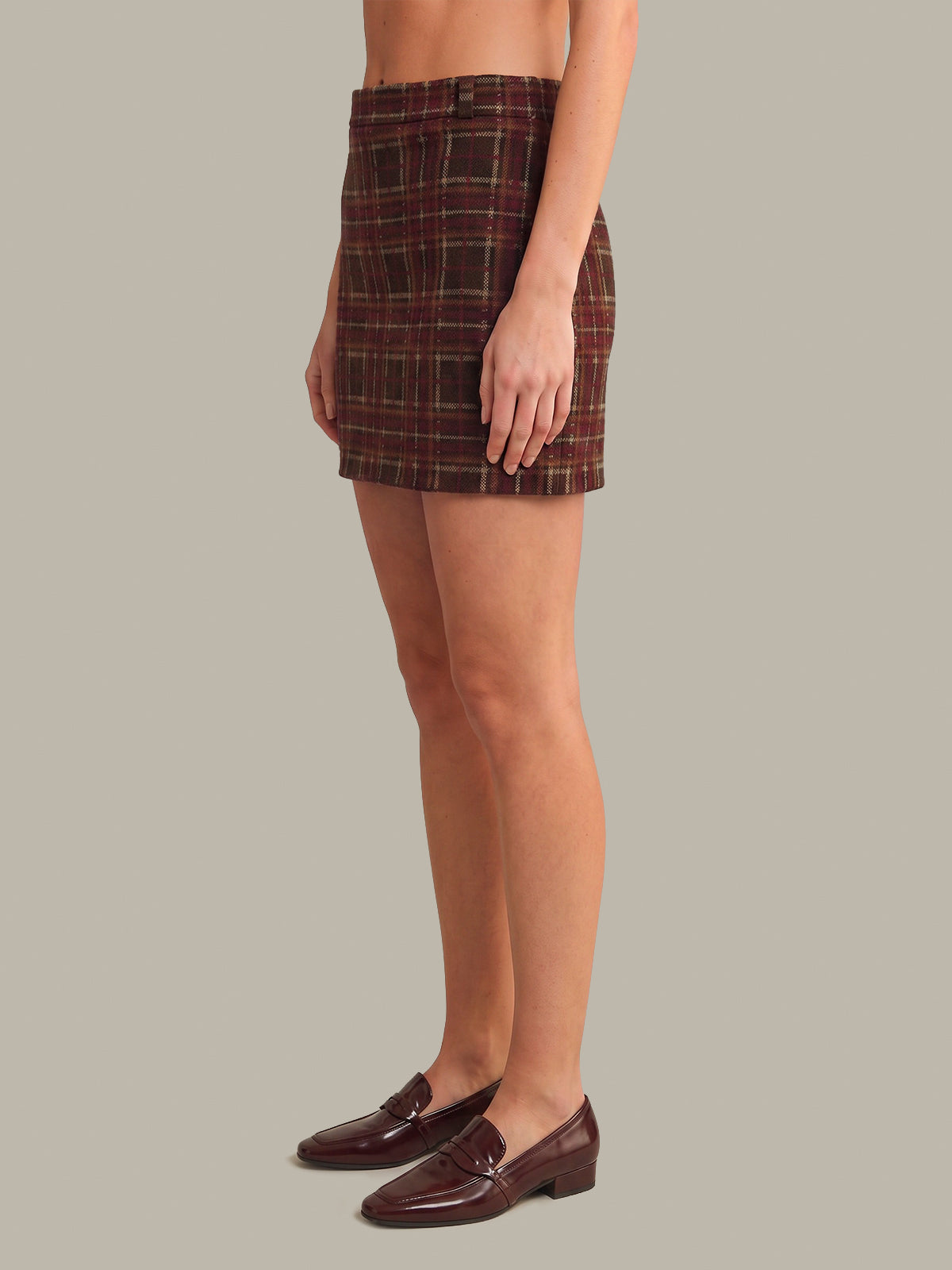 A model shown from the waist down wears a brown and red plaid mini skirt paired with dark brown leather penny loafers, set against a solid light gray background.
