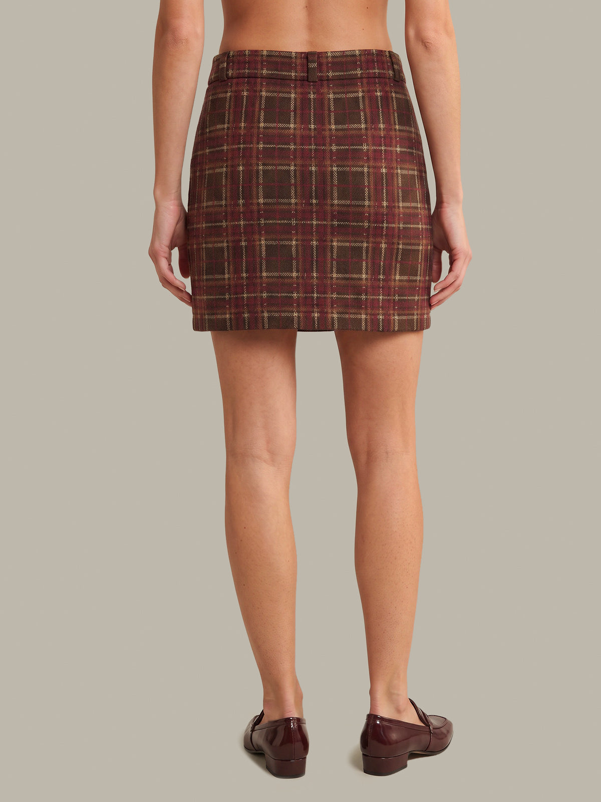 A model shown from the back wearing a brown and red plaid mini skirt with dark brown leather loafers, set against a solid light gray background.