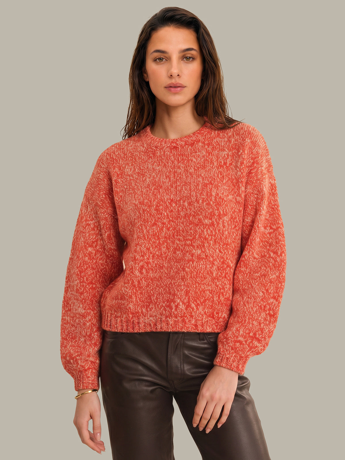 A woman wearing a textured, heathered coral-orange knit sweater with balloon sleeves and dark brown faux-leather pants, standing against a solid light gray background.