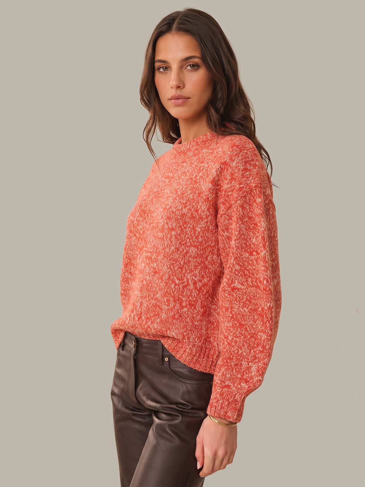 A woman wearing a textured, heathered coral-orange knit sweater and dark brown faux-leather pants, photographed against a solid light gray studio background.