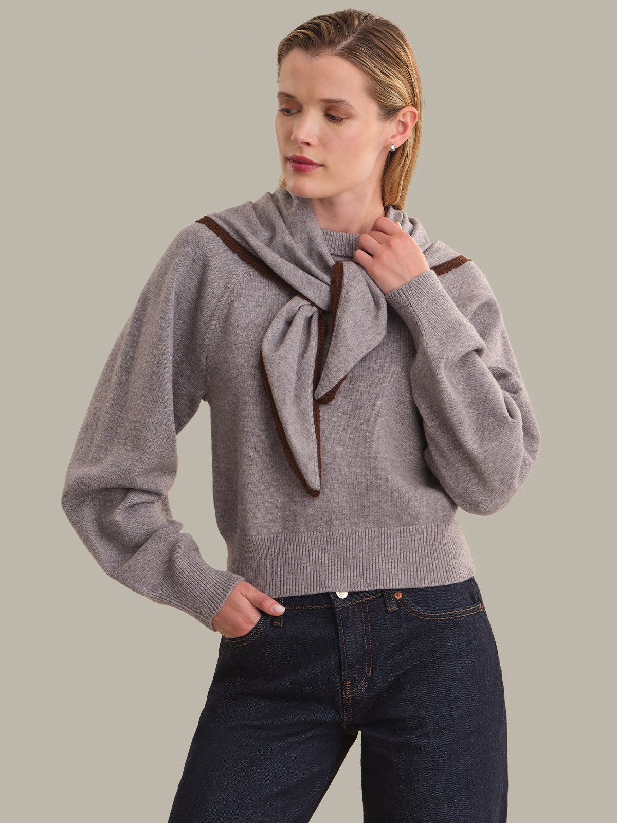 A woman wearing a light gray knit sweater with a large draped scarf detail and dark brown trim, paired with dark blue jeans, against a solid light gray background.